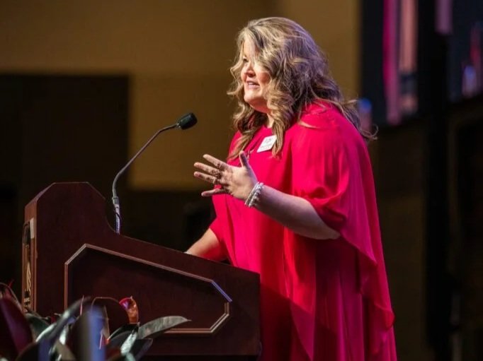 A woman with long, wavy gray hair, wearing a bright red dress, speaking at a podium with a microphone in a conference or event setting.
