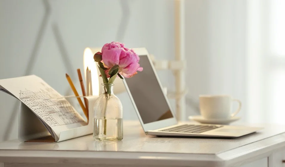 A white desk with a pink peony in a small glass bottle, an open notepad with pens, a laptop, a coffee cup, and a lit candle.
