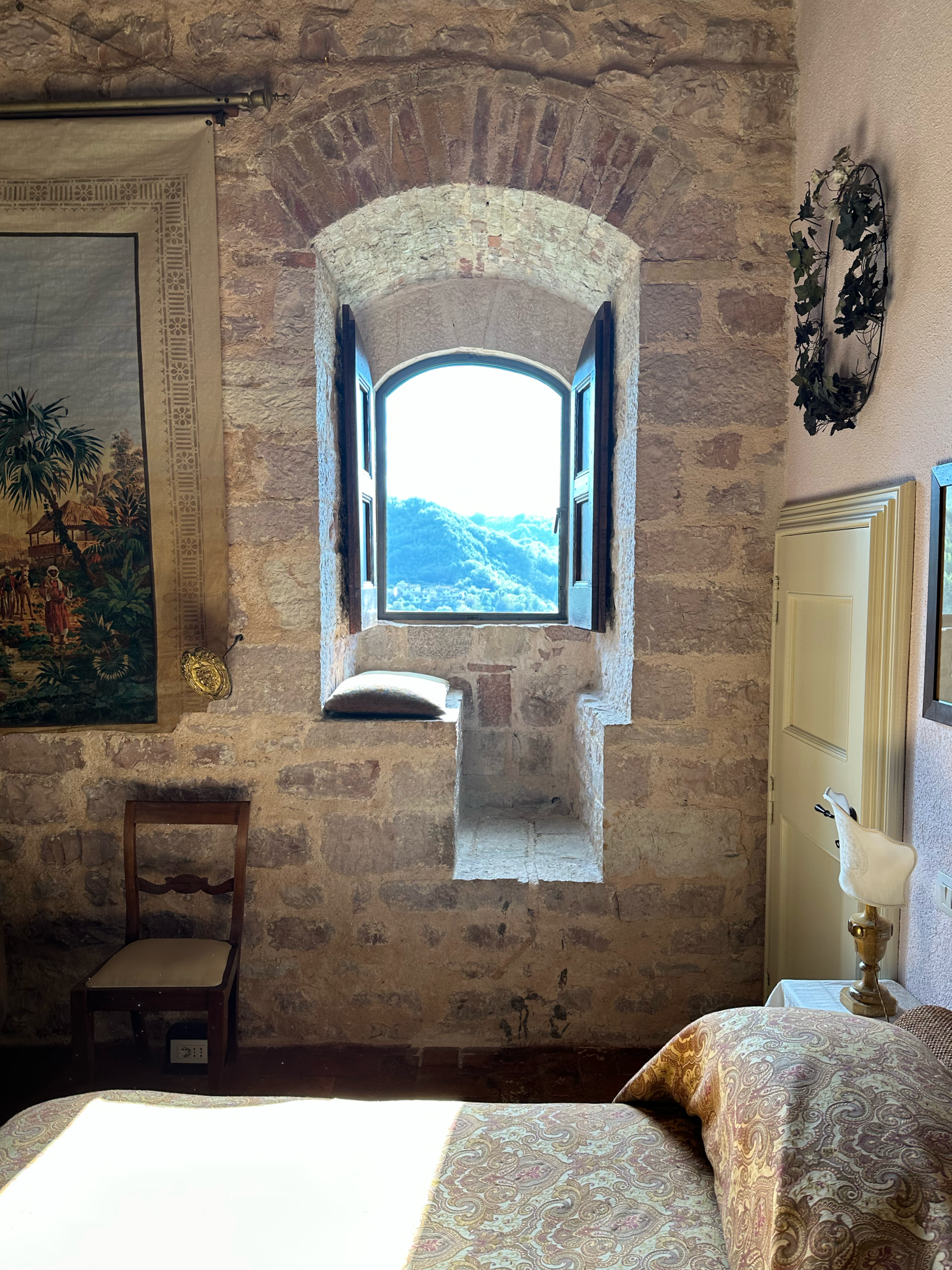 A room with a stone wall and an arched window showing a scenic view of mountains and sky. There is a wooden chair, a bed with ornate bedding, a nightstand with a lamp, and framed artwork on the wall.