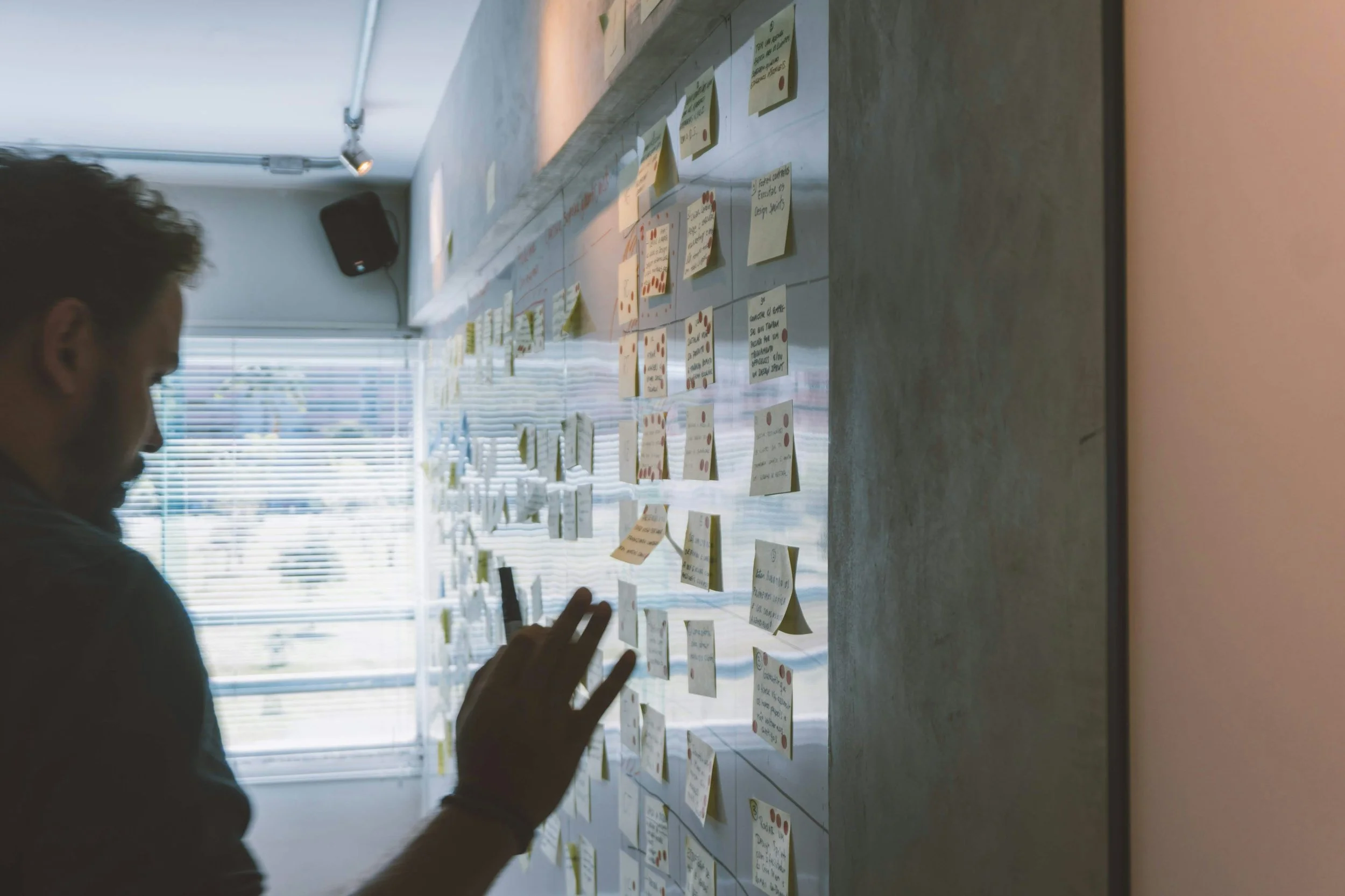 A person standing in front of a wall covered in numerous sticky notes, possibly organizing or analyzing ideas; sunlight coming through window blinds in the background.