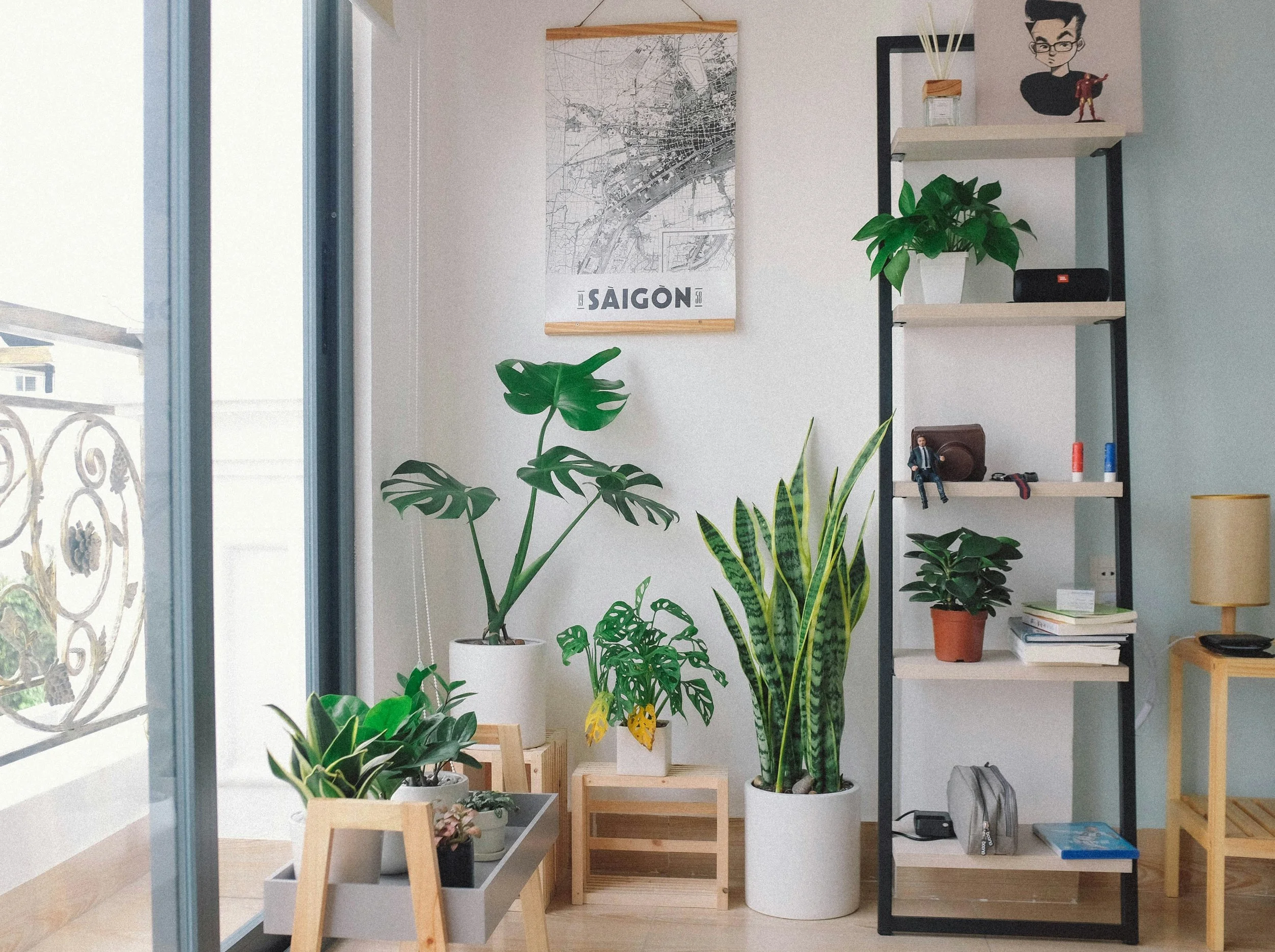 A well-lit interior space with several green houseplants, a black metal and wood shelf with decorative items, books, and small figurines, near a glass door with a balcony railing outside. A framed map of Saigon hangs on the wall.
