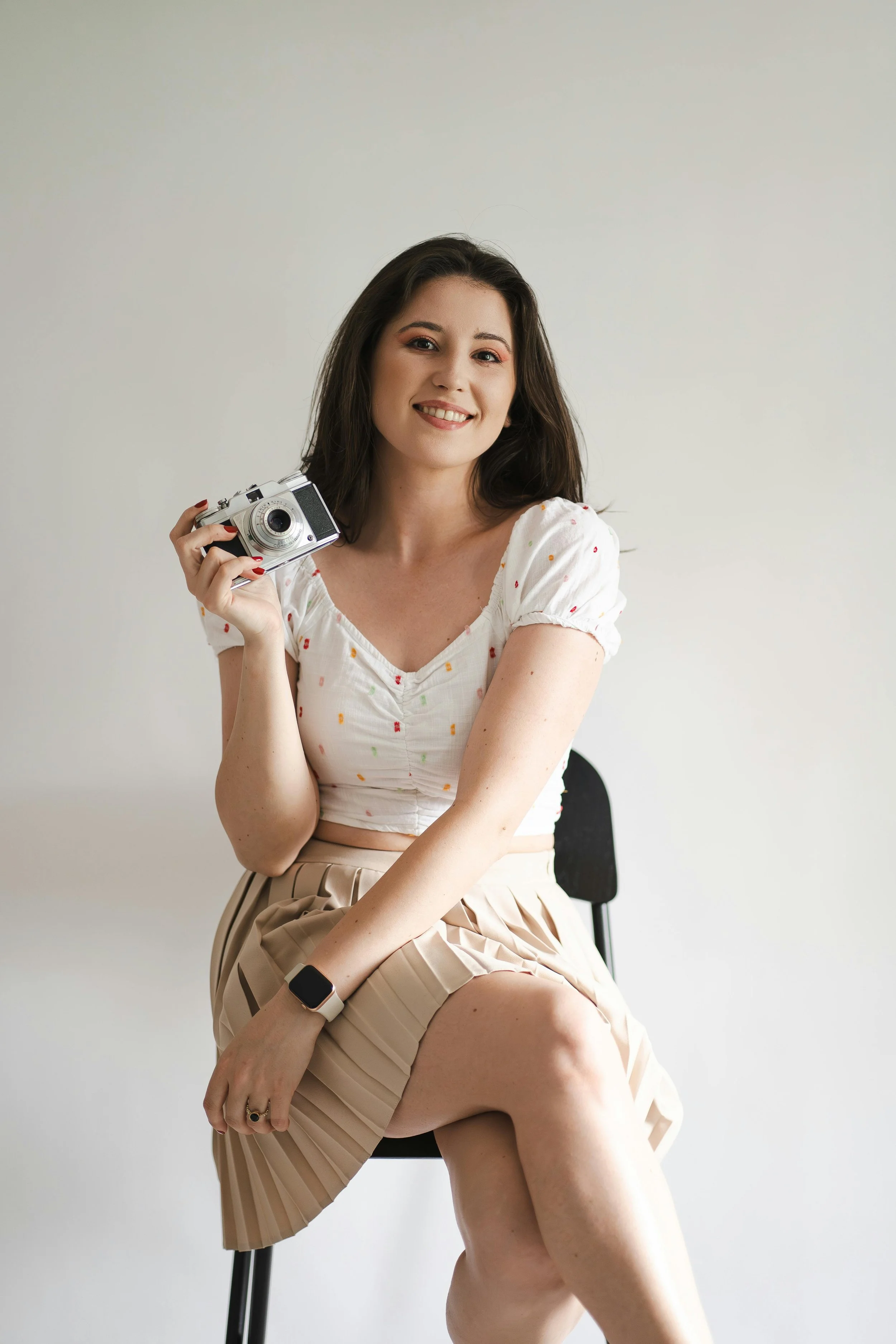 Young woman with dark hair smiling, seated on a black chair, holding a vintage camera in her right hand, wearing a white short-sleeve top with small colorful dots, a beige pleated skirt, and a smartwatch on her left wrist, against a plain light-colored background.