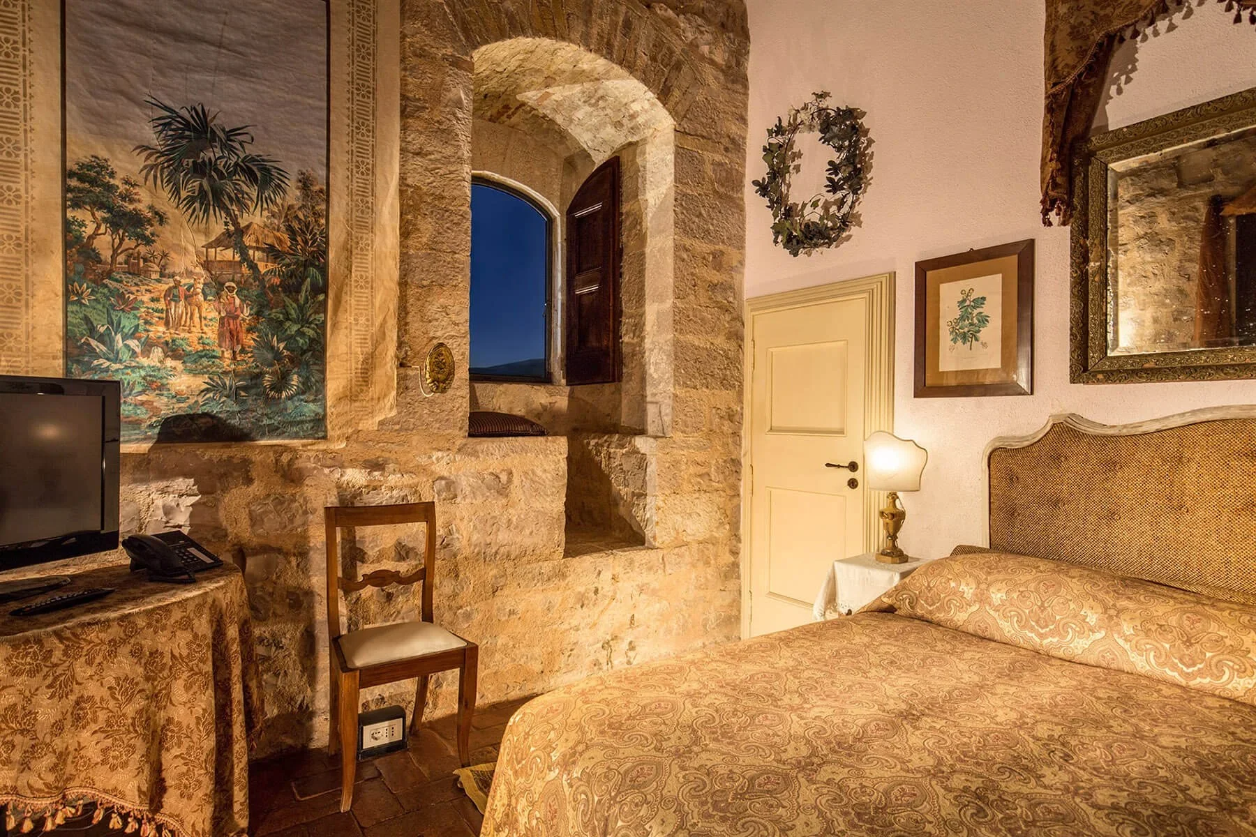 A cozy bedroom with a stone wall and an open window showing a night sky; a bed with a decorative headboard and patterned bedding; framed artwork and a wreath on the wall; a lamp on a side table; a small wooden chair; a table with a tablecloth and a TV; warm lighting creates a rustic ambiance.