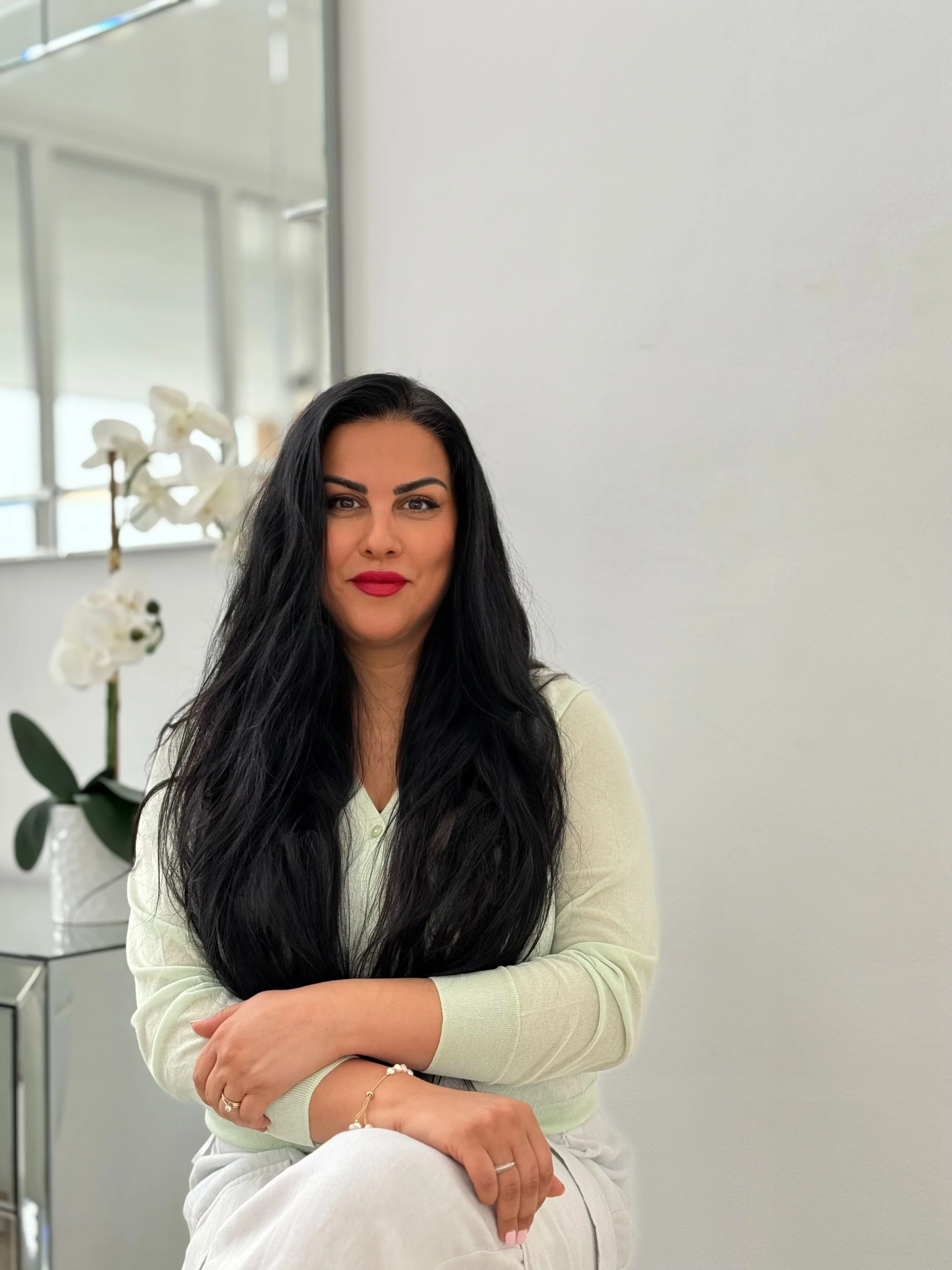 A woman with long black hair and red lipstick sitting indoors, next to a white orchid in a vase, with a mirror and window in the background.