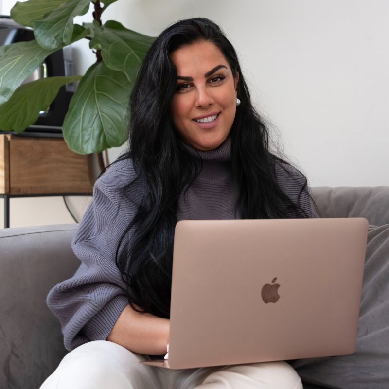 A woman with long black hair, smiling, sitting on a gray couch with a rose gold MacBook on her lap. She is wearing a gray turtleneck sweater and white pants, with a large green plant and a wooden cabinet in the background.