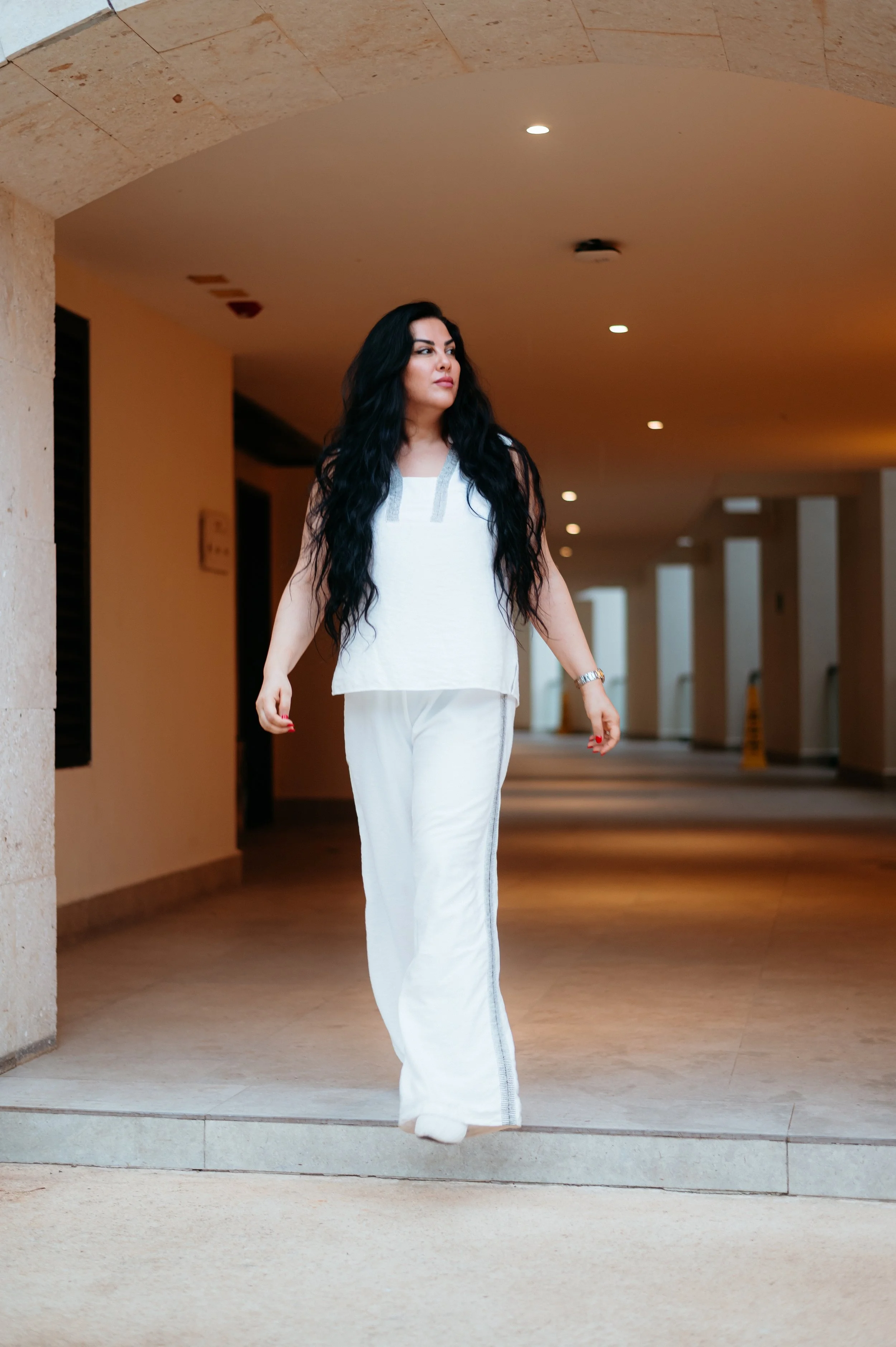 A woman with long black hair, wearing a white sleeveless top and white pants, walks through a lobby or hallway with beige walls and ceiling, with ceiling lights and a caution wet floor sign in the background.