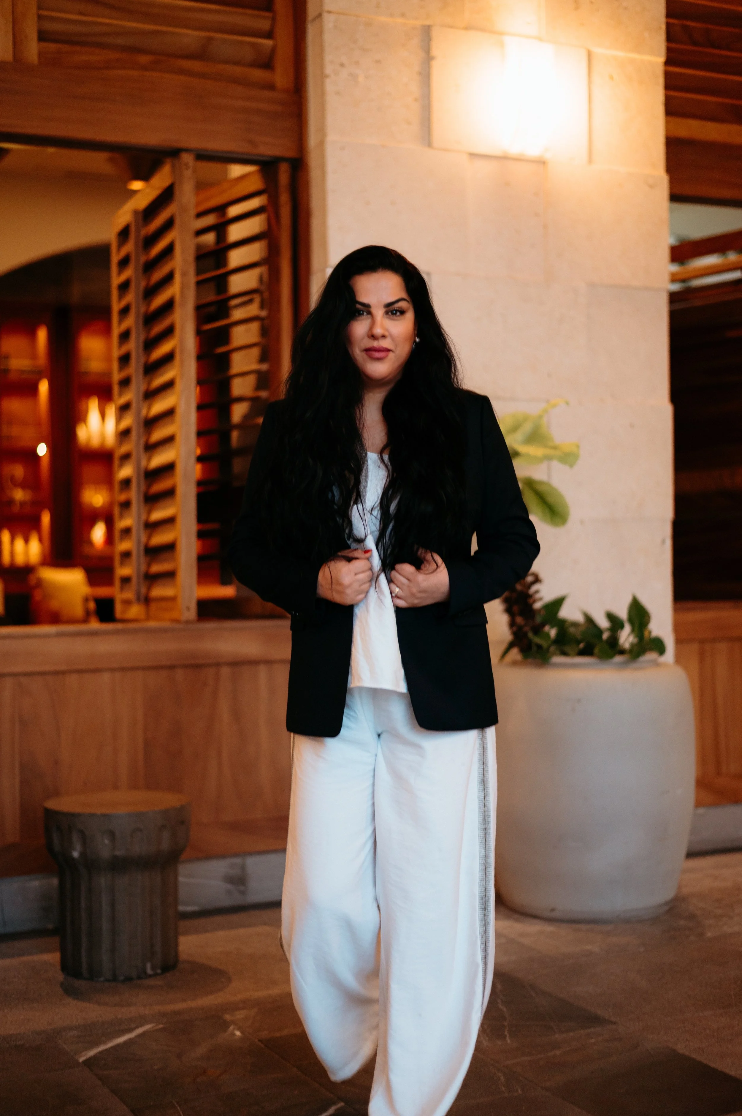 Woman with long black hair, wearing a black blazer and white pants, standing indoors with wooden decor and plants in the background.