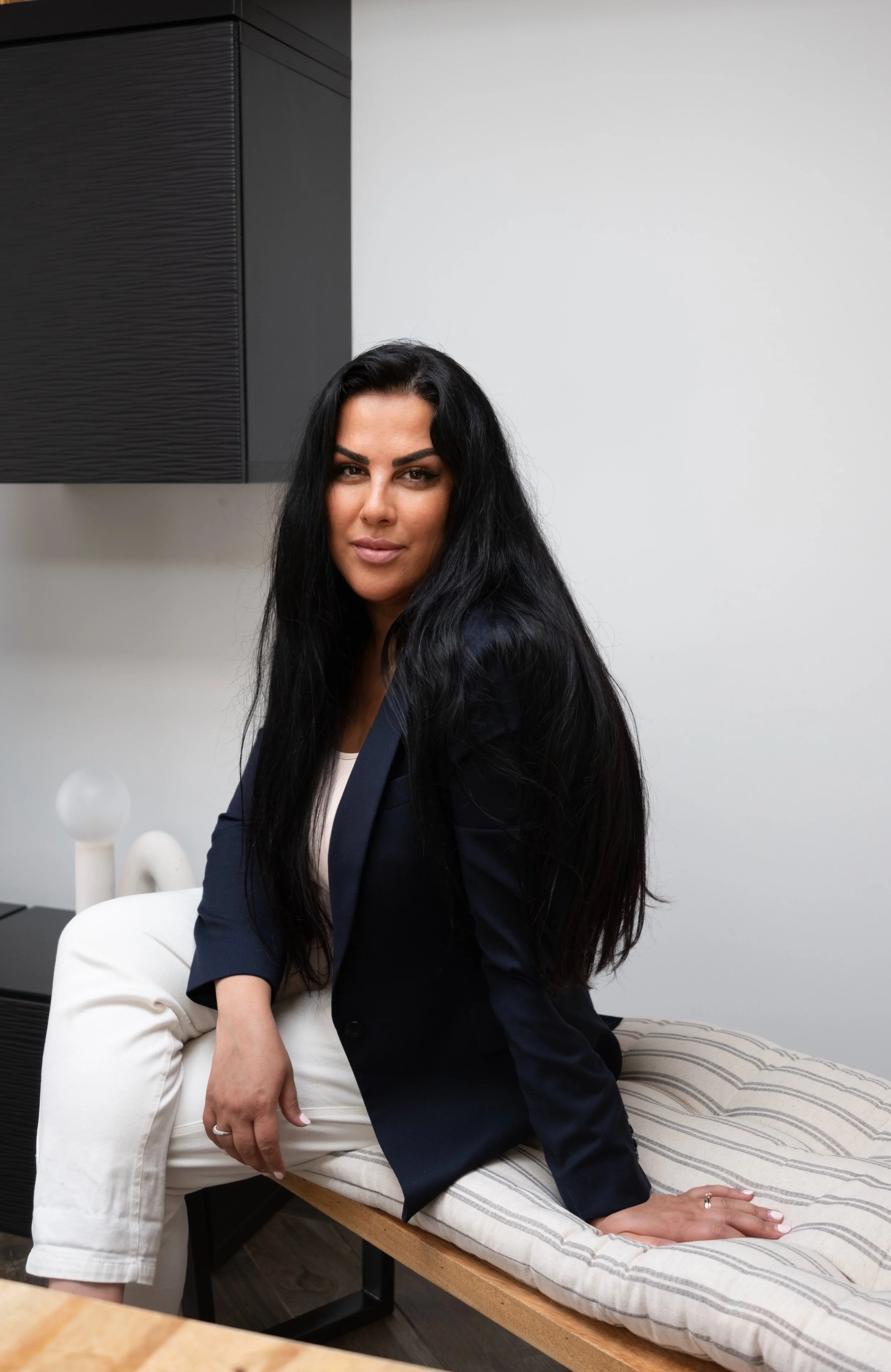 A woman with long black hair sitting on a striped cushion on a wooden bench, wearing a dark blazer and white pants, in a modern interior setting.