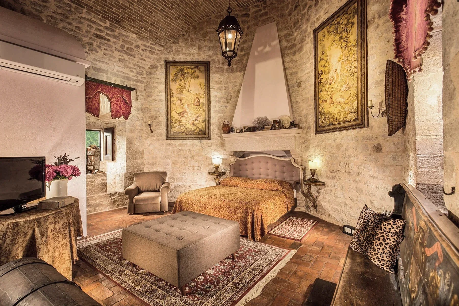 A cozy bedroom with stone walls, a brick ceiling, and warm lighting. There is a bed with a luxurious headboard, side tables with lamps, and framed artwork on the walls. A small armchair, a patterned rug, and various decorative items complete the room's vintage and rustic decor.