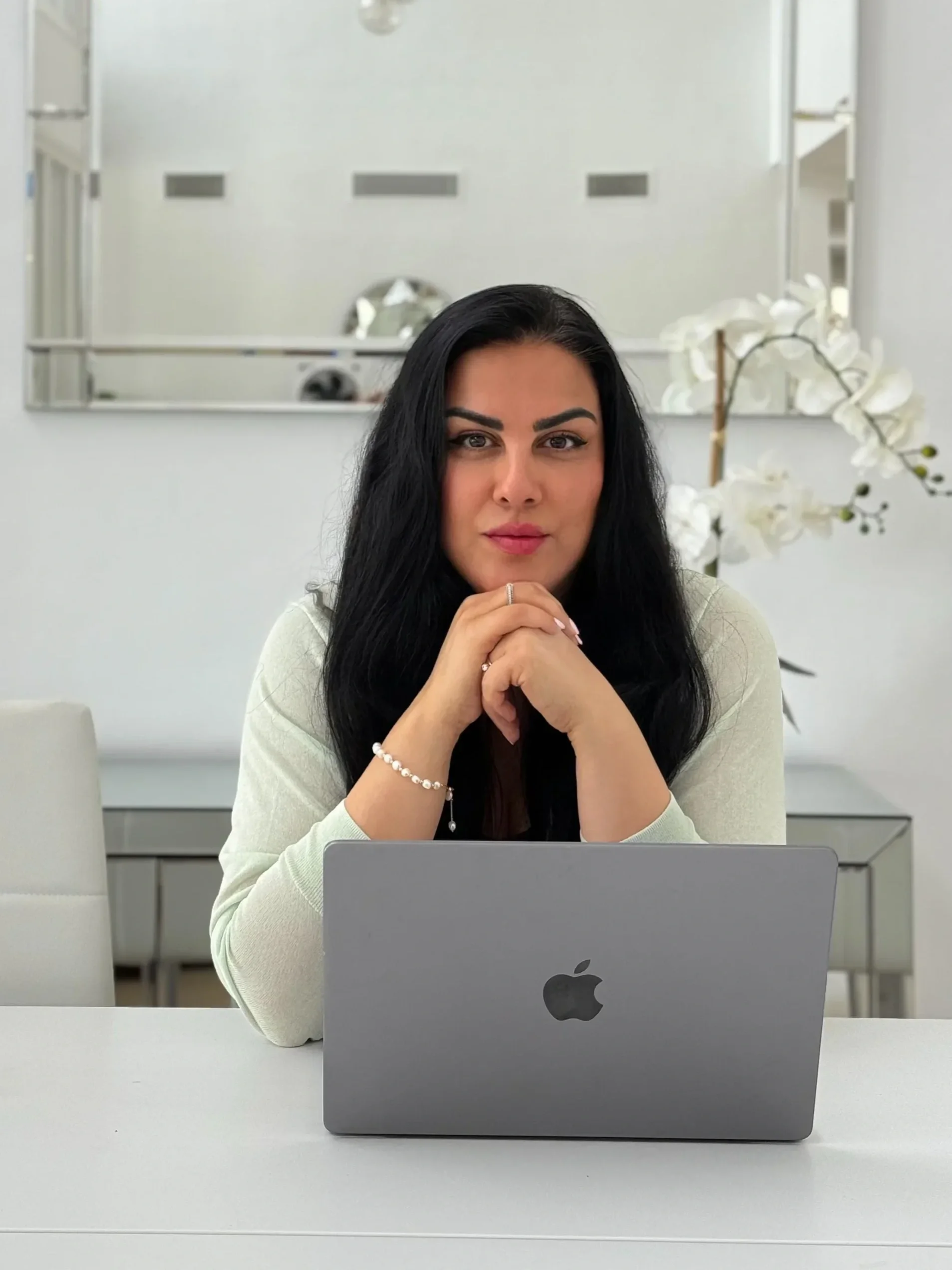 A woman with long black hair sitting at a desk with a silver MacBook, hands clasped under her chin, in a bright office space with white walls and decorative white orchids in the background.