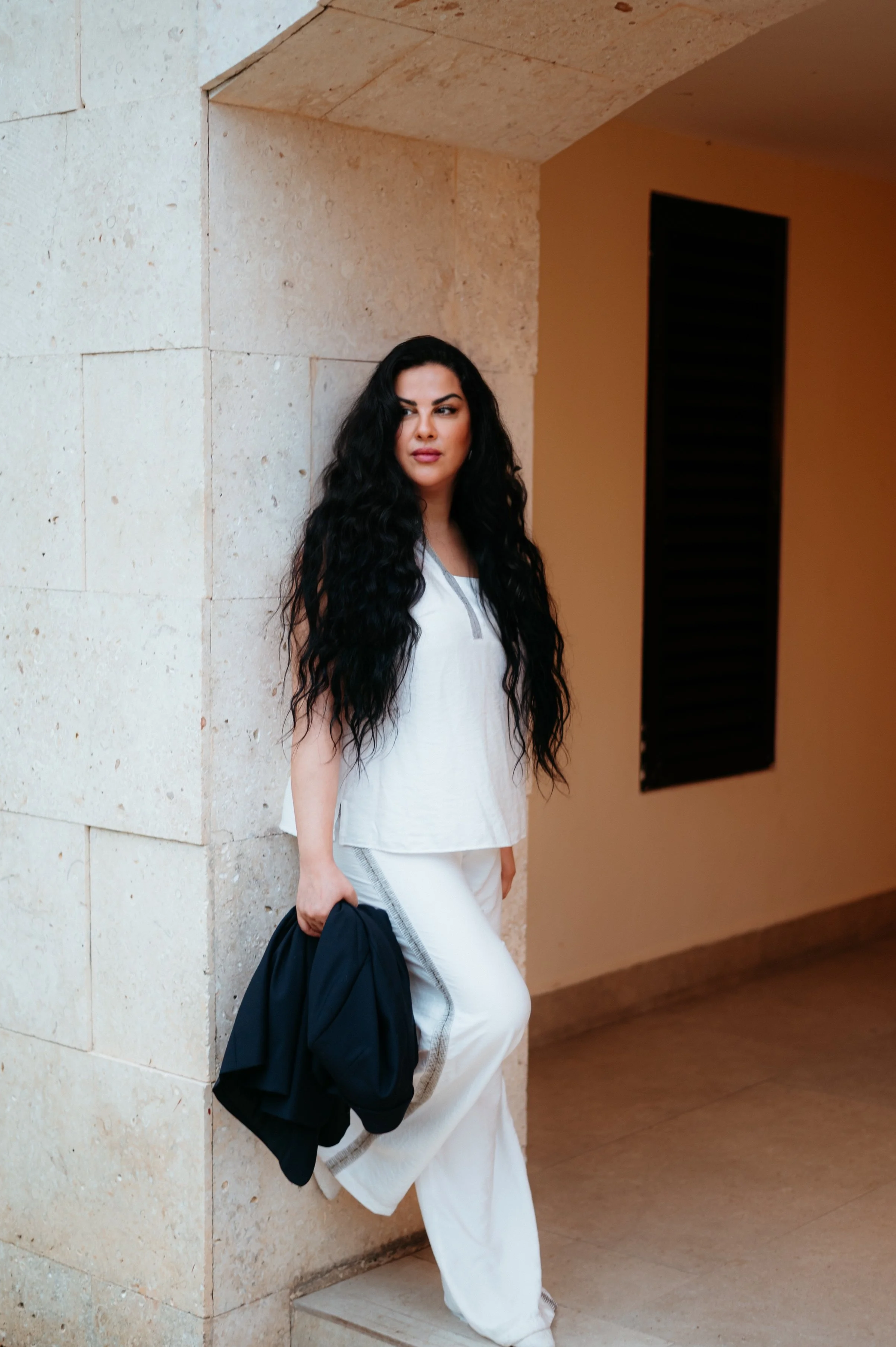 A woman with long, wavy black hair wearing a white top and white pants, holding a black jacket, leaning against a beige stone wall in an indoor corridor.
