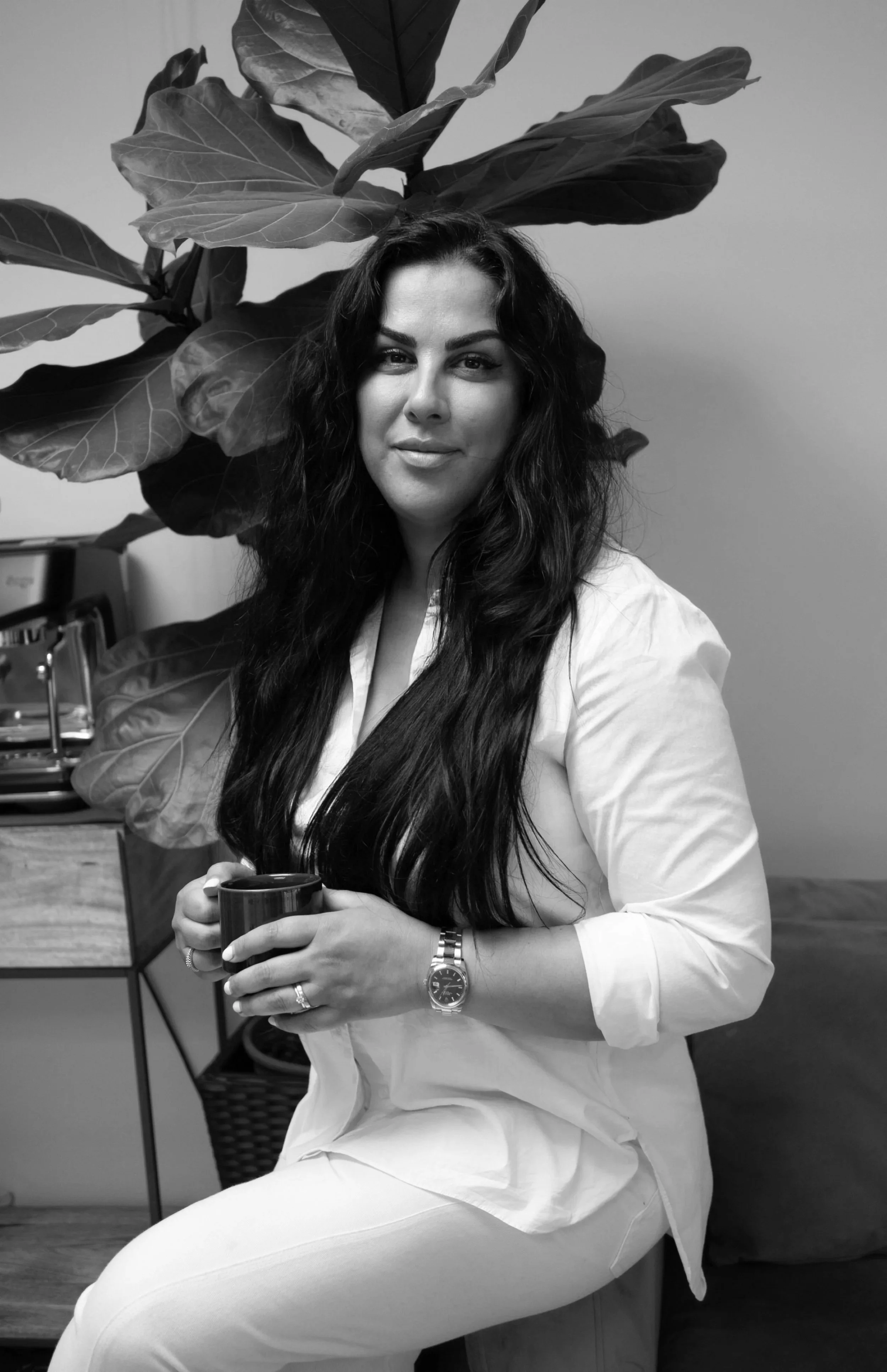 A woman with long dark hair, wearing a white shirt and light-colored pants, sitting on a chair and holding a mug, with a large leafy plant in the background.