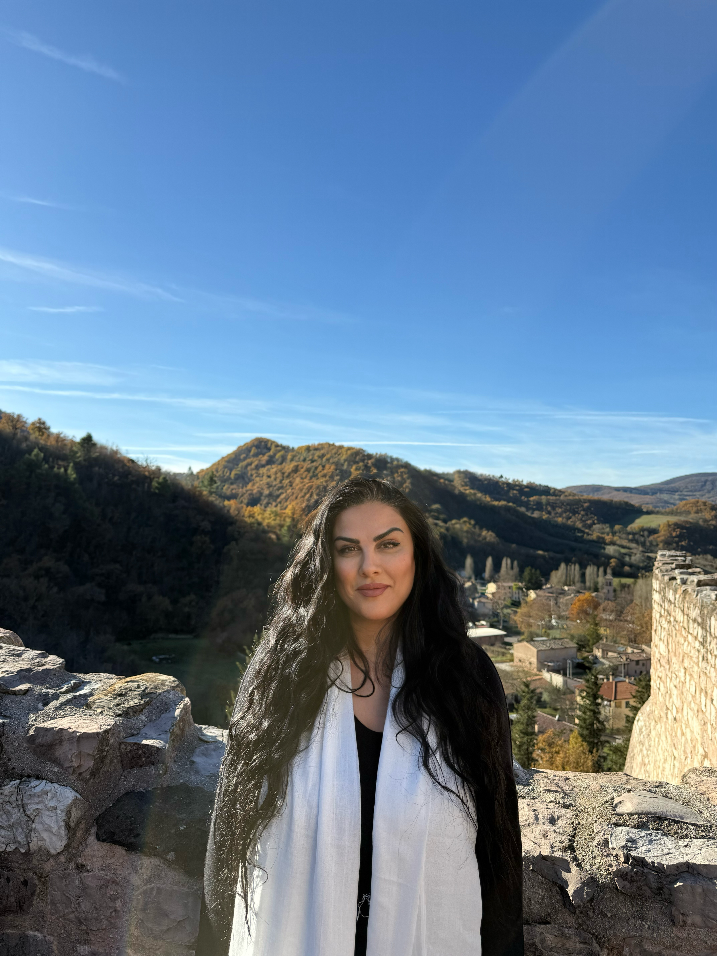 A woman with long dark curly hair standing outdoors with mountains and a small town in the background under a clear blue sky.