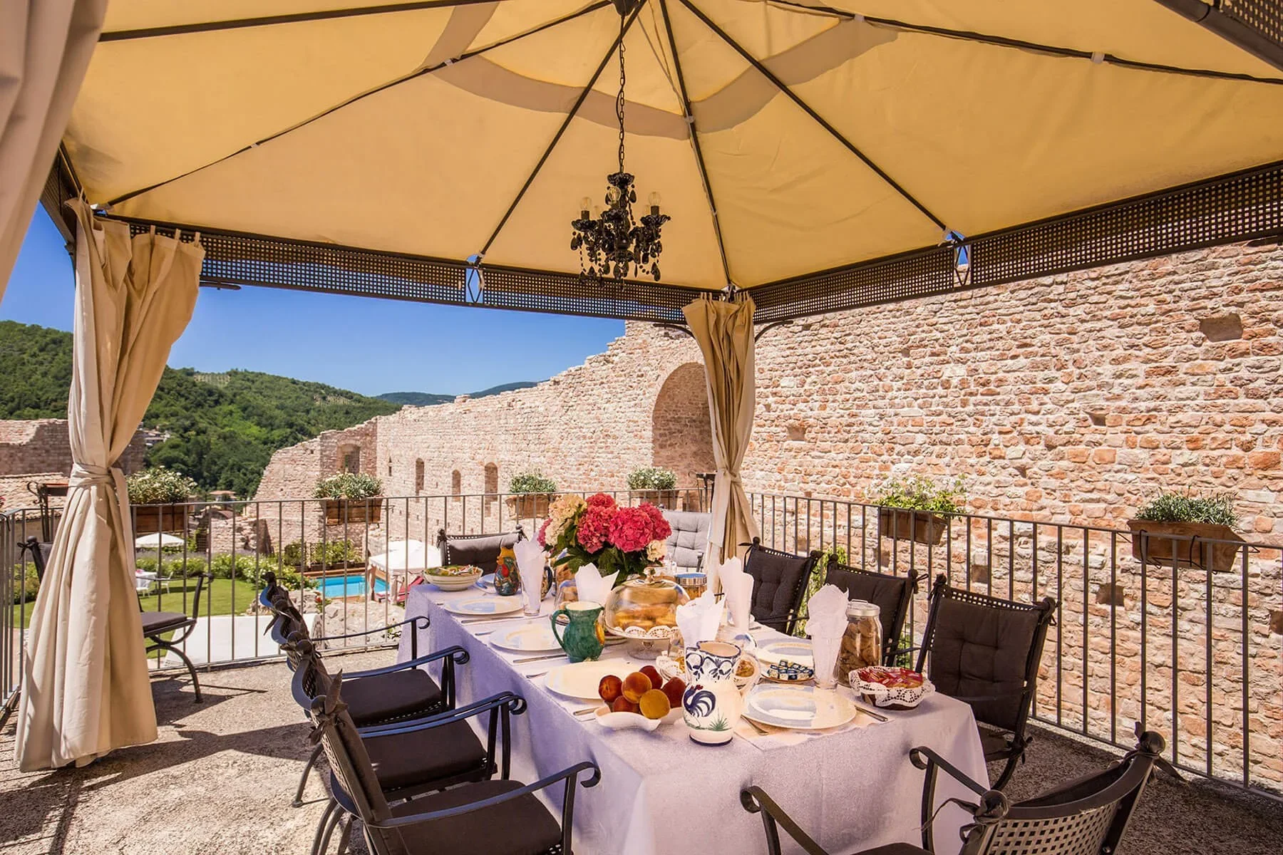Outdoor dining area under a large beige canopy with a black chandelier hanging from the center. The table is set with white tablecloth, dishes, a flower centerpiece, and food. The scene includes a brick wall, potted plants on the railing, and a view of a green landscape and blue sky in the background.