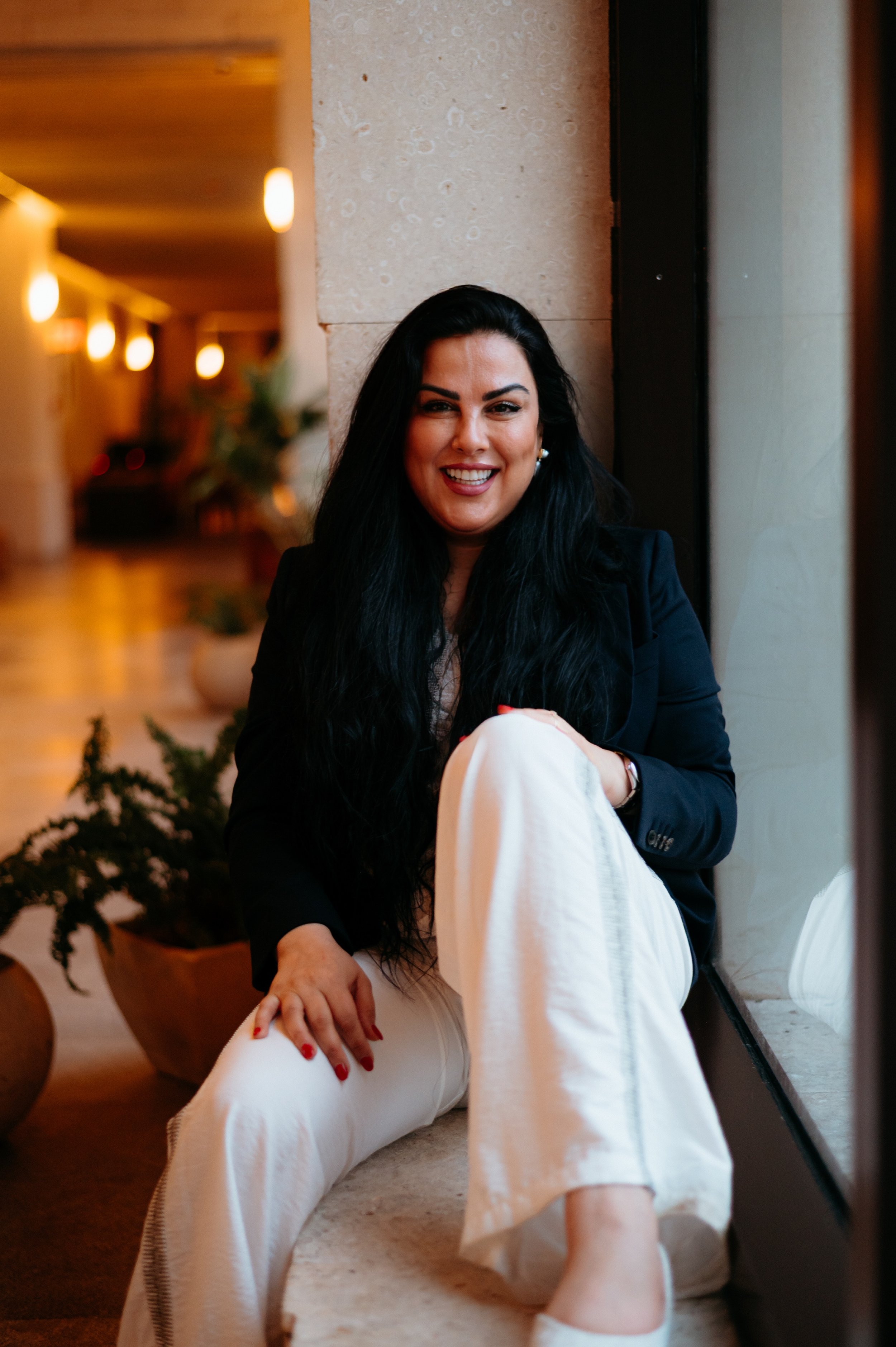 A woman with long black hair, wearing a black blazer and white pants, sitting by a window in a warmly lit indoor setting, smiling at the camera.