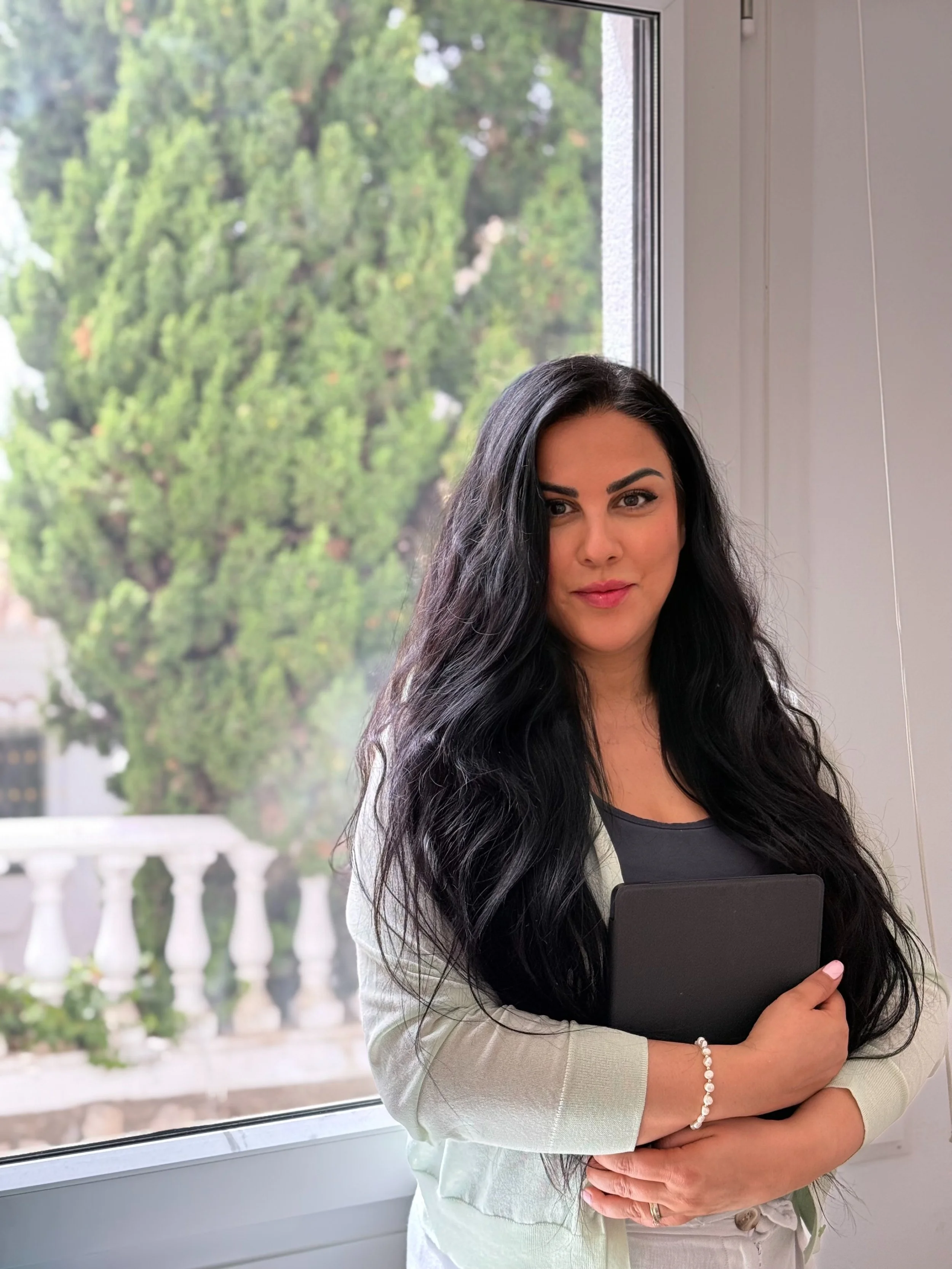 A woman with long black hair holding a tablet in front of a window with trees outside.