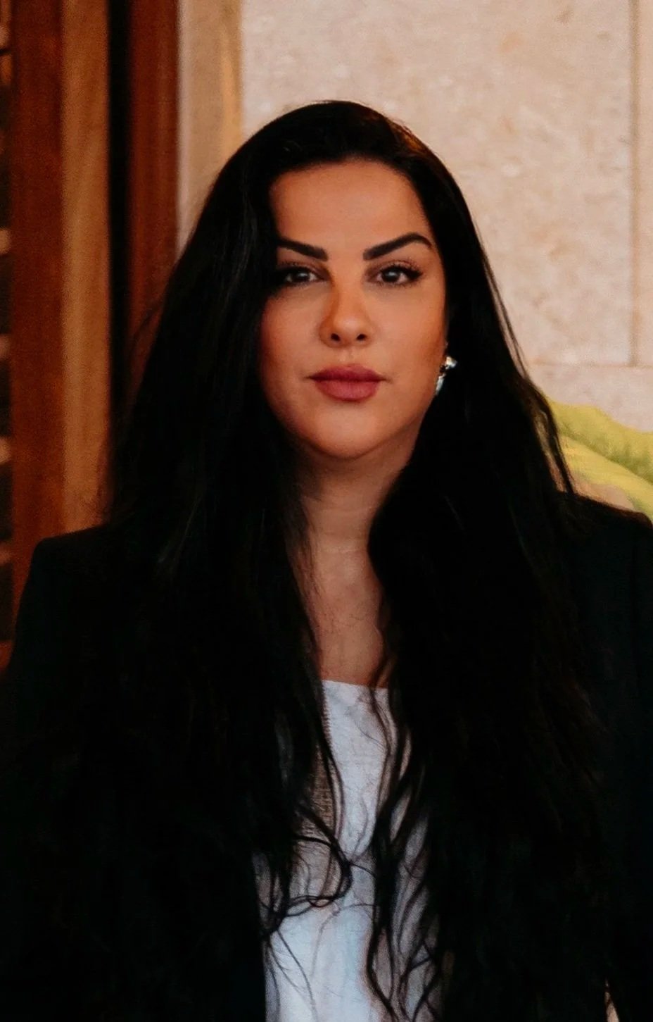 A woman with long black hair and light skin, wearing a black jacket and white top, posing indoors with a neutral background.