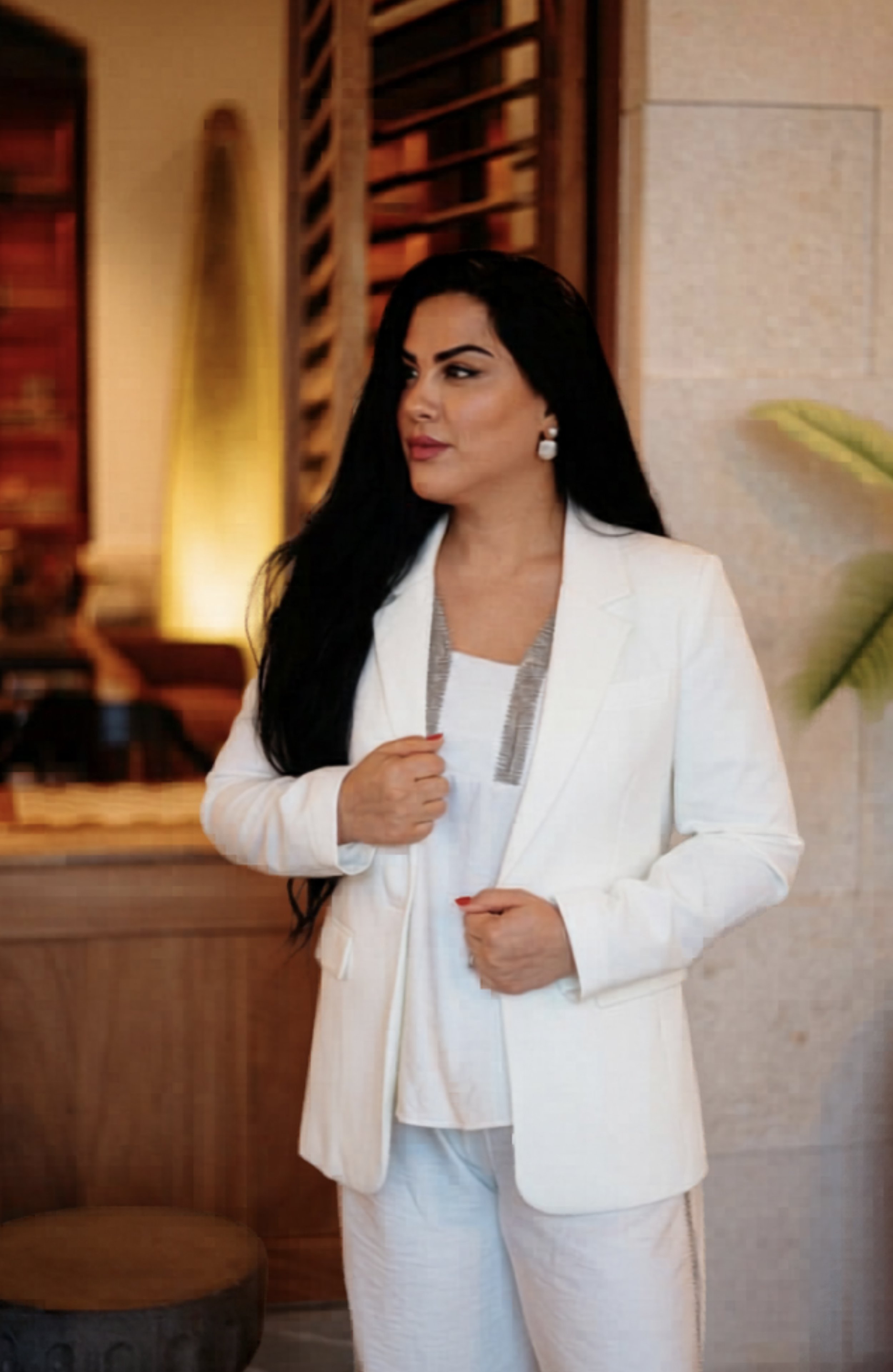 A woman with long black hair wearing a white suit and jewelry standing indoors with a background of warm lighting, wooden decor, and a large green plant.
