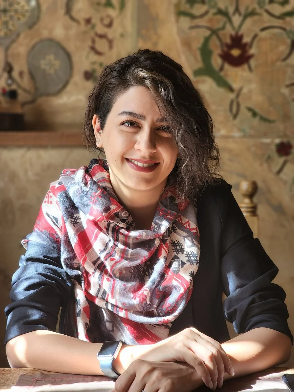 A woman with short dark and gray hair, wearing a black shirt and a colorful patterned scarf, smiling while sitting at a table with a wooden background and decorative art.
