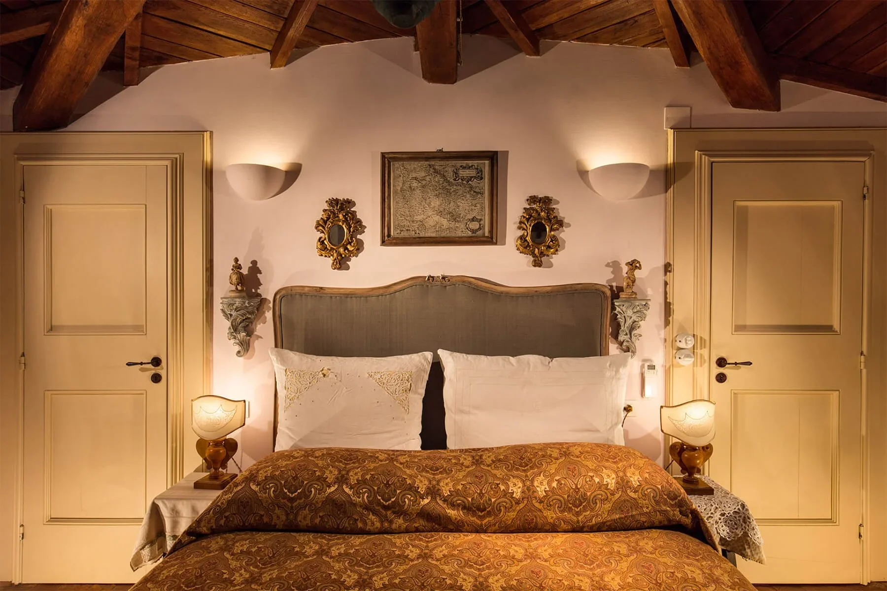 A cozy bedroom with twin beds, ornate headboards, and vintage decor. The wall behind the beds has framed artwork and decorative mirrors, with two wall-mounted lights. There are matching nightstands with lamps on each side of the beds, a patterned bedspread, and closed doors on both sides of the room. The ceiling features exposed wooden beams.