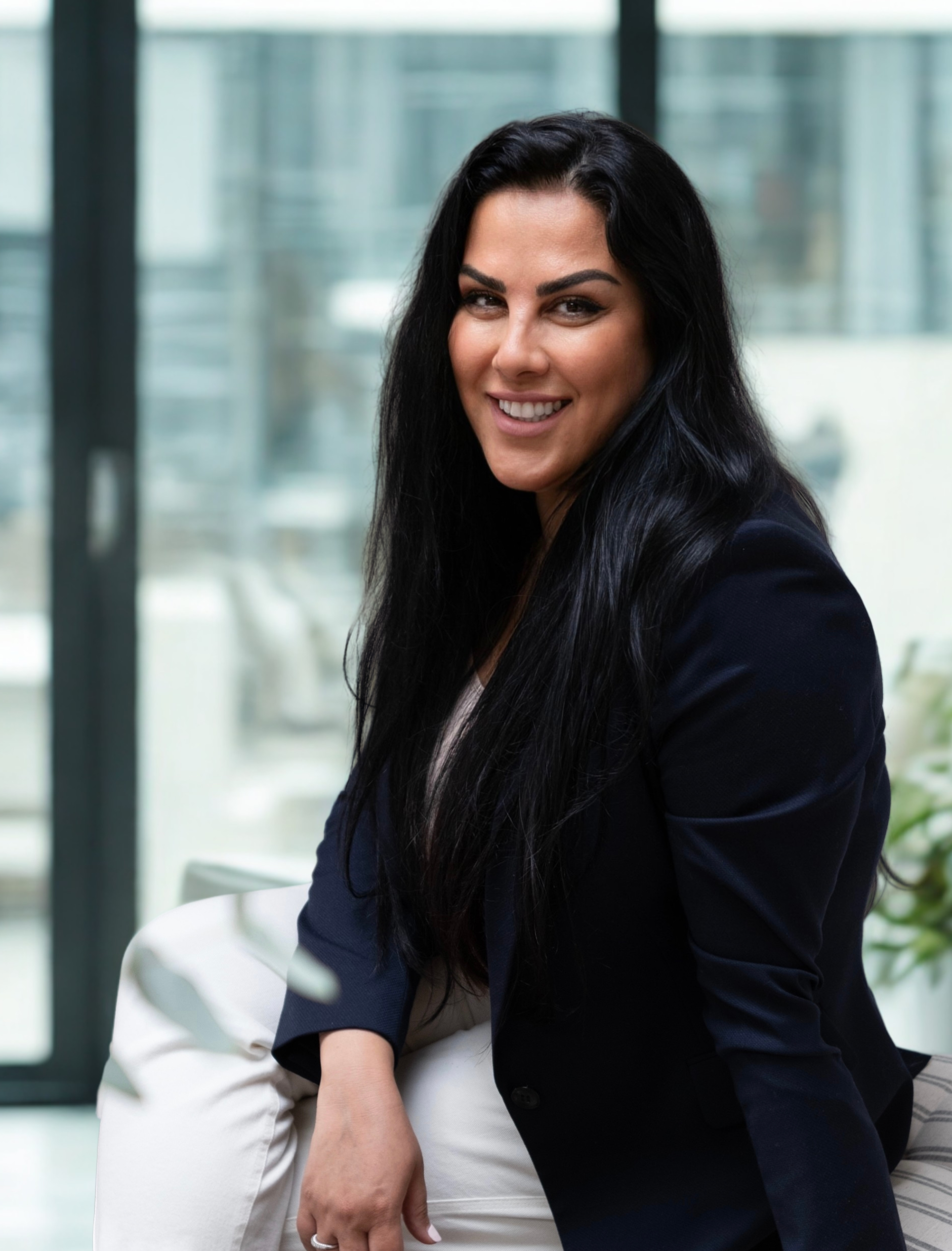 A woman with long black hair smiling and sitting indoors near large windows.