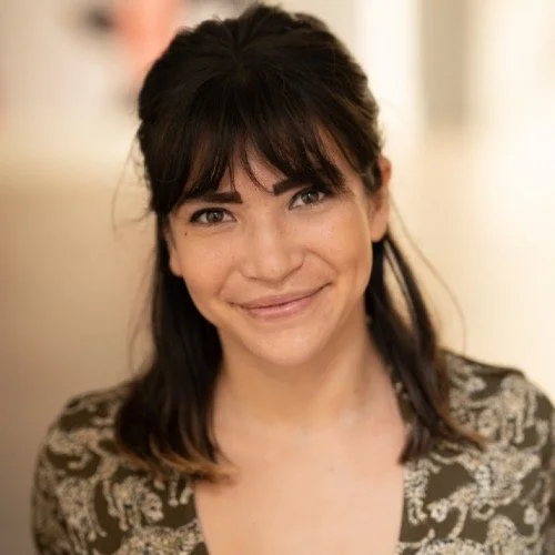 A woman with dark hair and bangs smiling at the camera, wearing a patterned top.