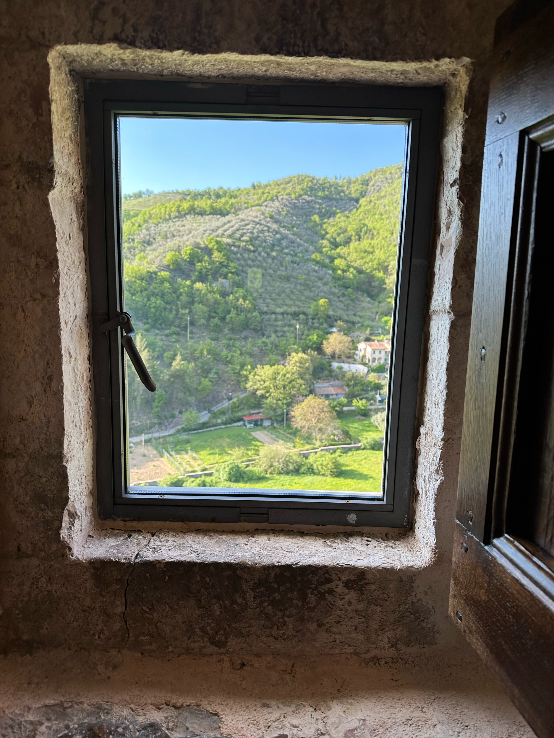 View through a small window showing a lush green hillside with trees and a few houses below under a clear blue sky.