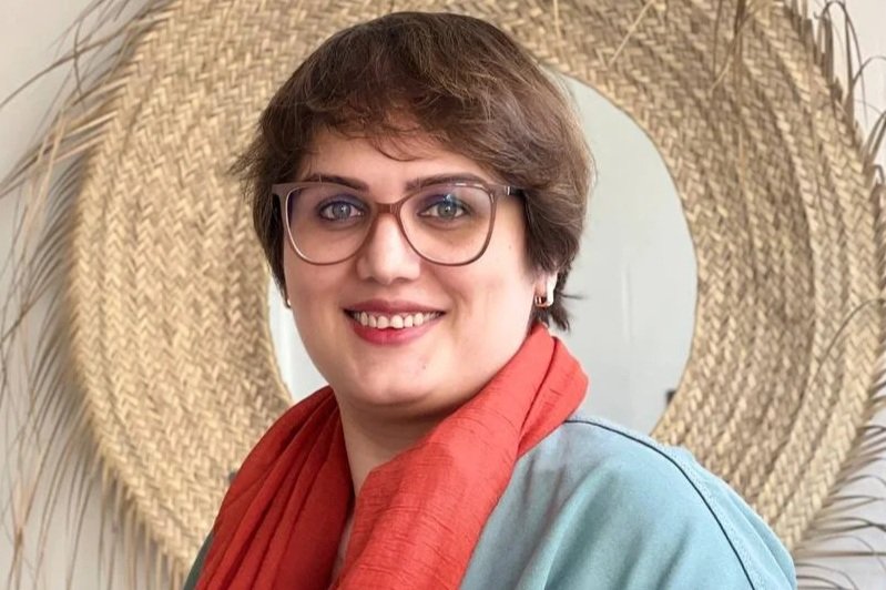 A woman with short brown hair, wearing glasses, earrings, a blue shirt, and an orange scarf, smiling at the camera with a woven circular wall hanging behind her.