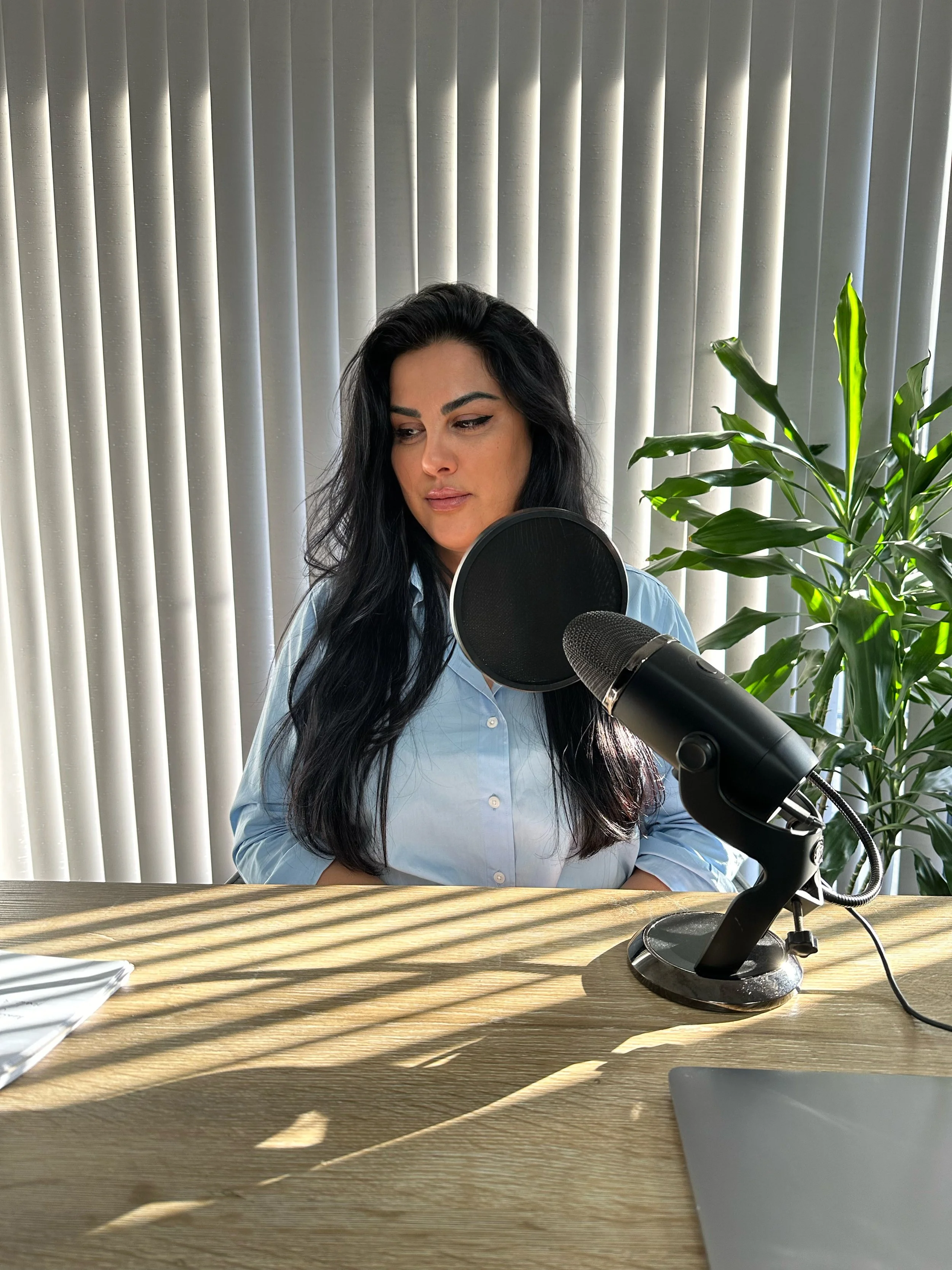 A woman with long black hair, wearing a light blue shirt, sitting at a wooden desk in front of a microphone with a pop filter, in a room with vertical blinds and a green plant.