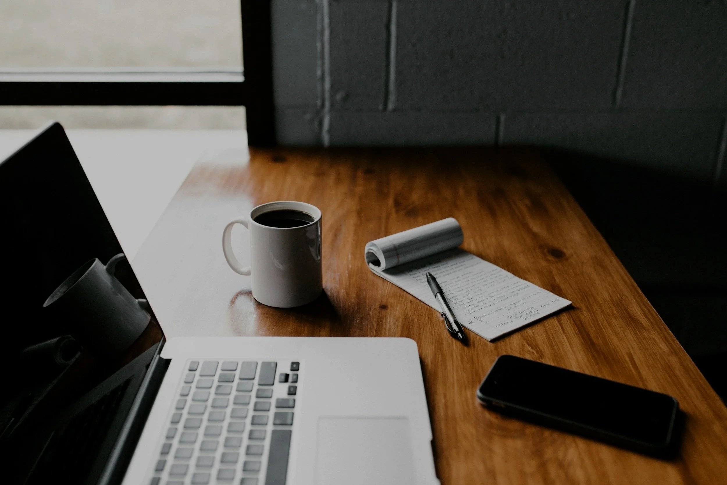 A wooden desk with a laptop, a white cup of black coffee, a notepad with a pen, and a smartphone, near a window with blinds.