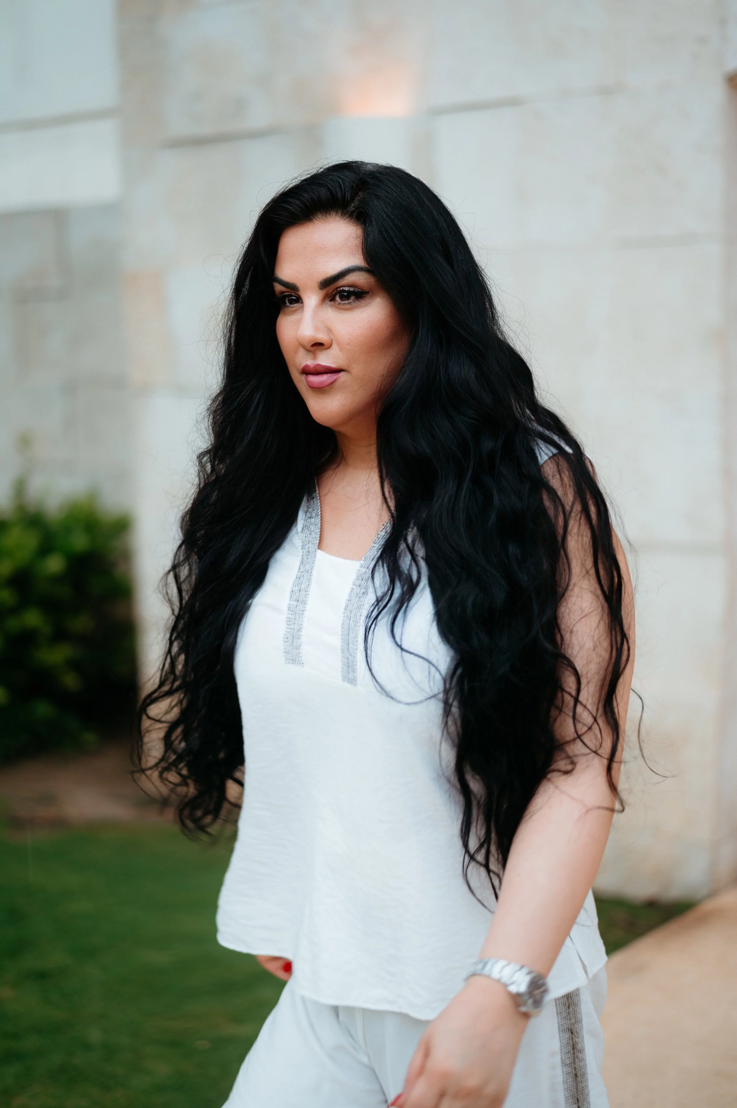A woman with long black wavy hair wearing a sleeveless white top and white pants, standing outdoors with a stone wall and greenery in the background.