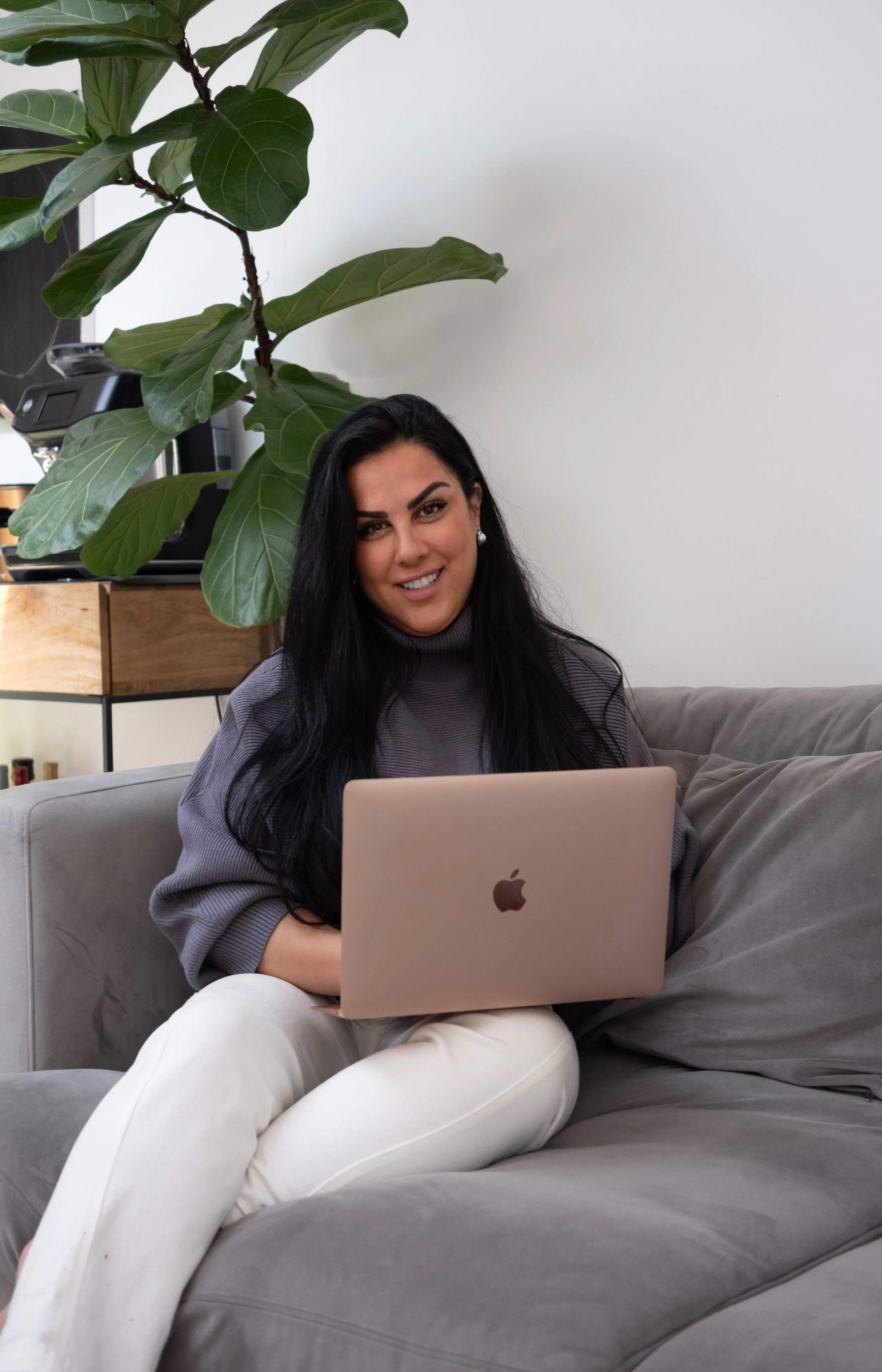 A woman with long black hair sitting on a gray couch, smiling while using an Apple MacBook laptop, with a large green plant and a wooden shelf in the background.