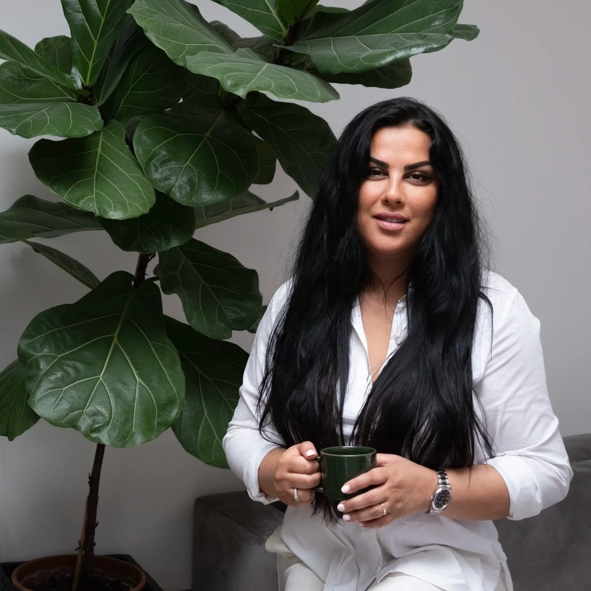 A woman with long black hair and wearing a white shirt holding a green mug, standing next to a large green leafy plant against a plain wall.
