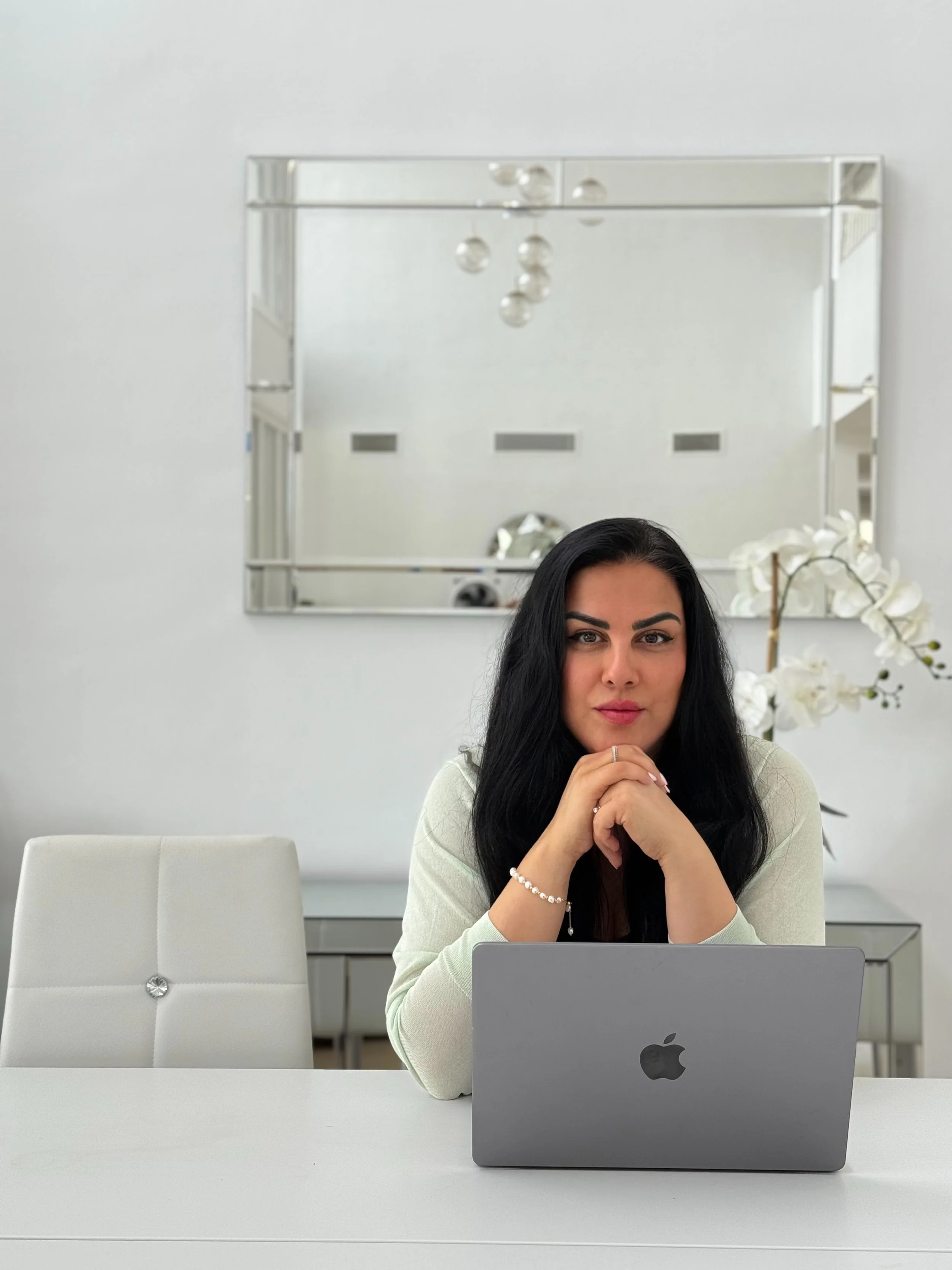 Woman with long black hair sitting at a white desk, with a gray Apple laptop in front of her, in a bright minimalistic room with a white chair and a mirror on the wall.