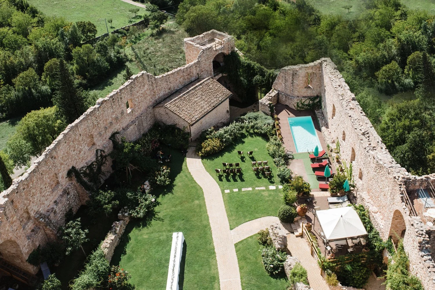 Aerial view of a courtyard with lush green grass, a swimming pool with blue water, outdoor seating, and surrounded by ancient stone walls, with trees and greenery in the background.
