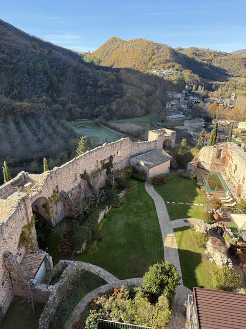 Aerial view of ancient stone ruins with arches, a garden, and a swimming pool, surrounded by green hills and a small town in the background.