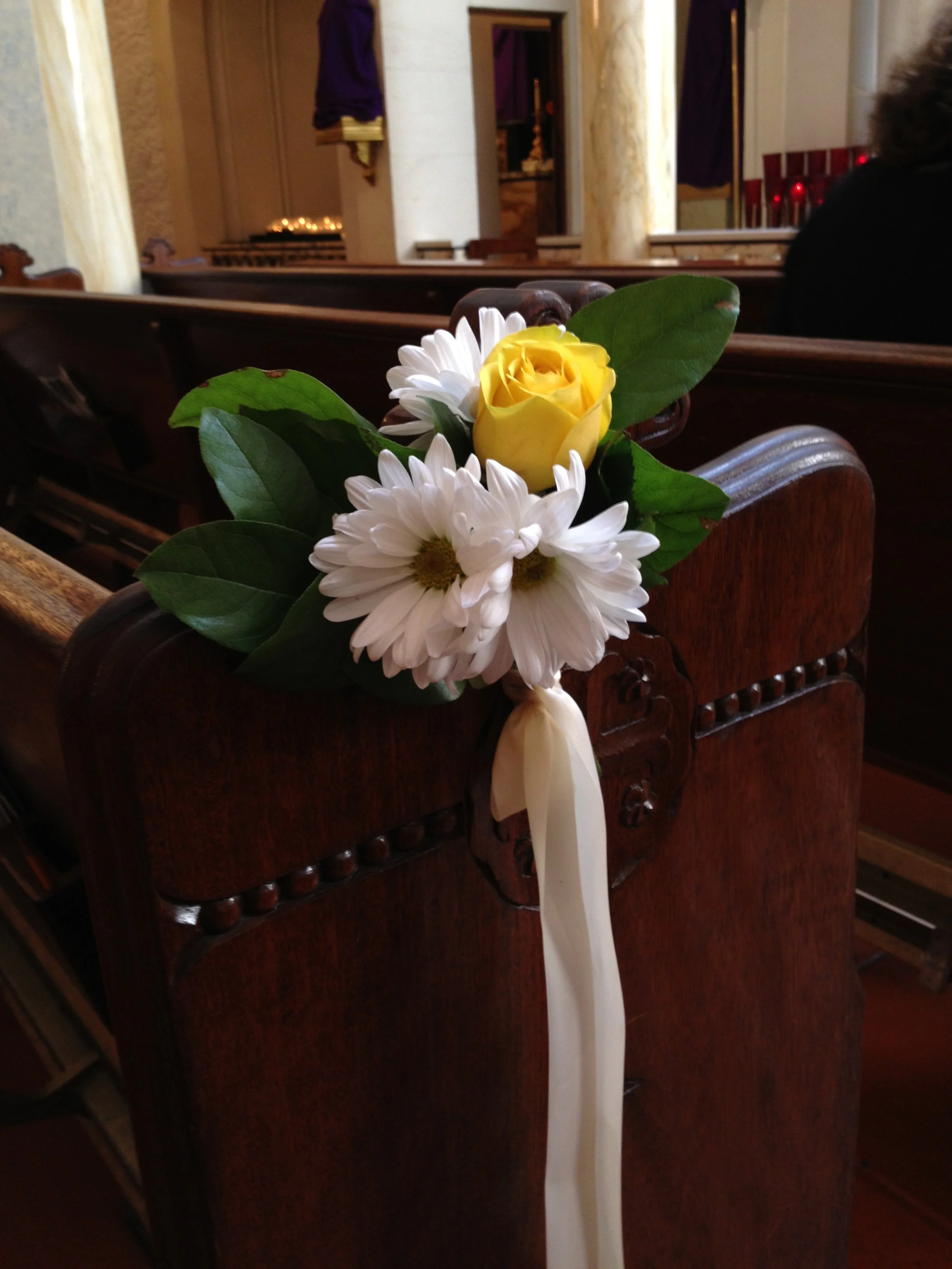 Pew-end flowers in a church: a yellow rose, white daisies, and greenery tied with cream ribbon