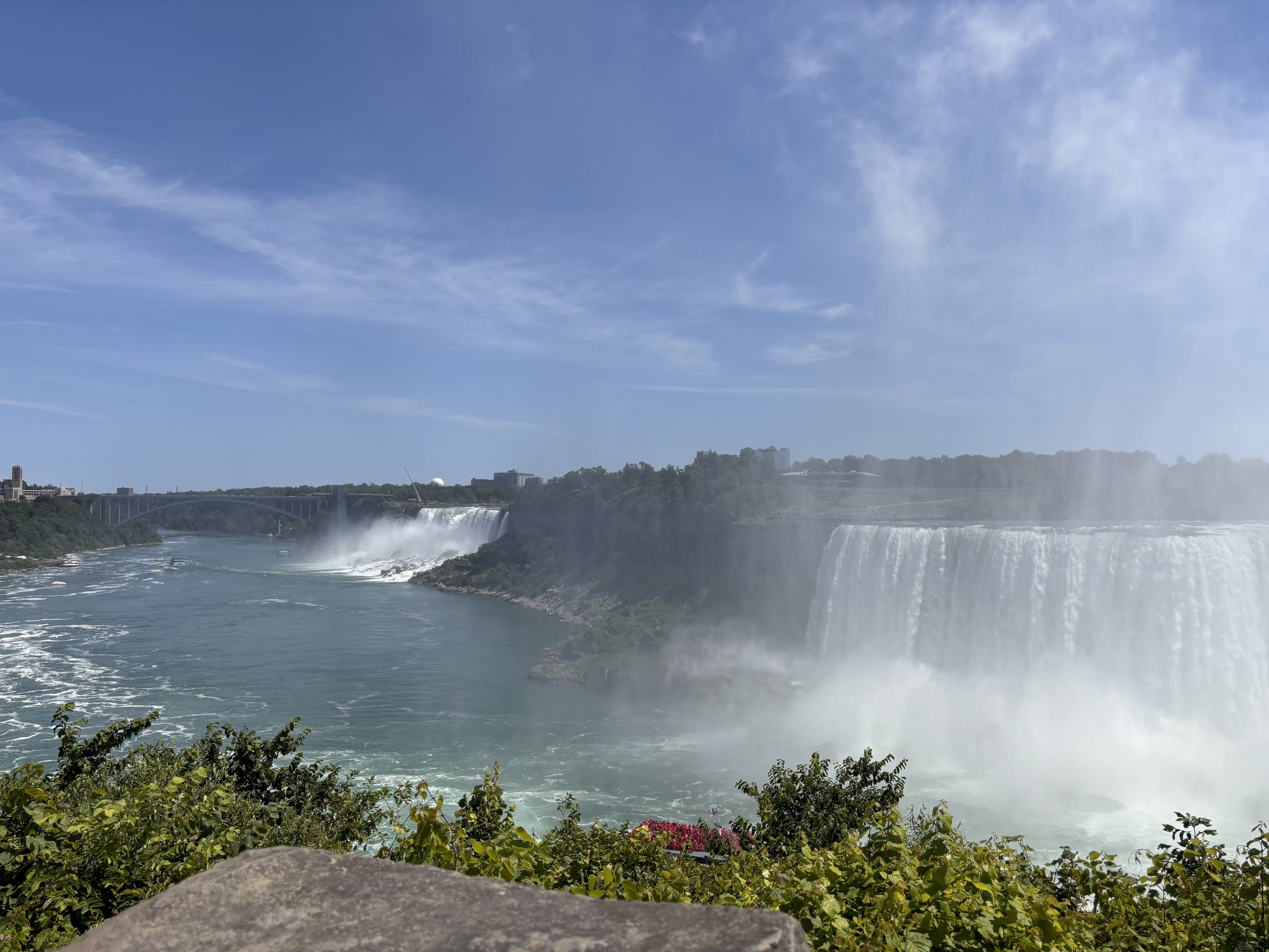 Niagara Falls with cascading waterfalls, mist rising, blue sky and some clouds, greenery in the foreground, and bridges in the distance.
