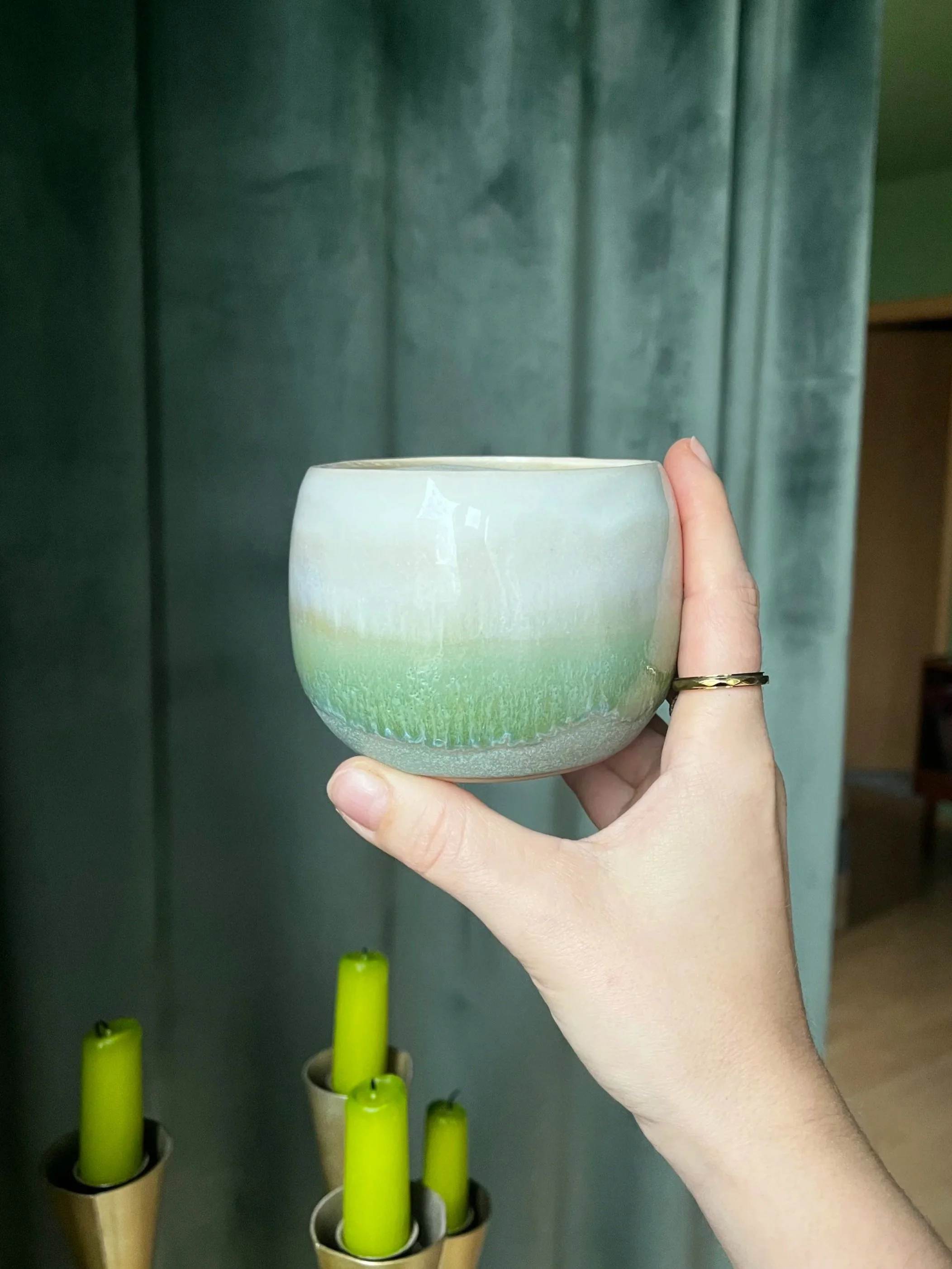 Hand hält eine Keramikschale mit grünem und cremefarbenem Muster vor einem grünen Vorhang.