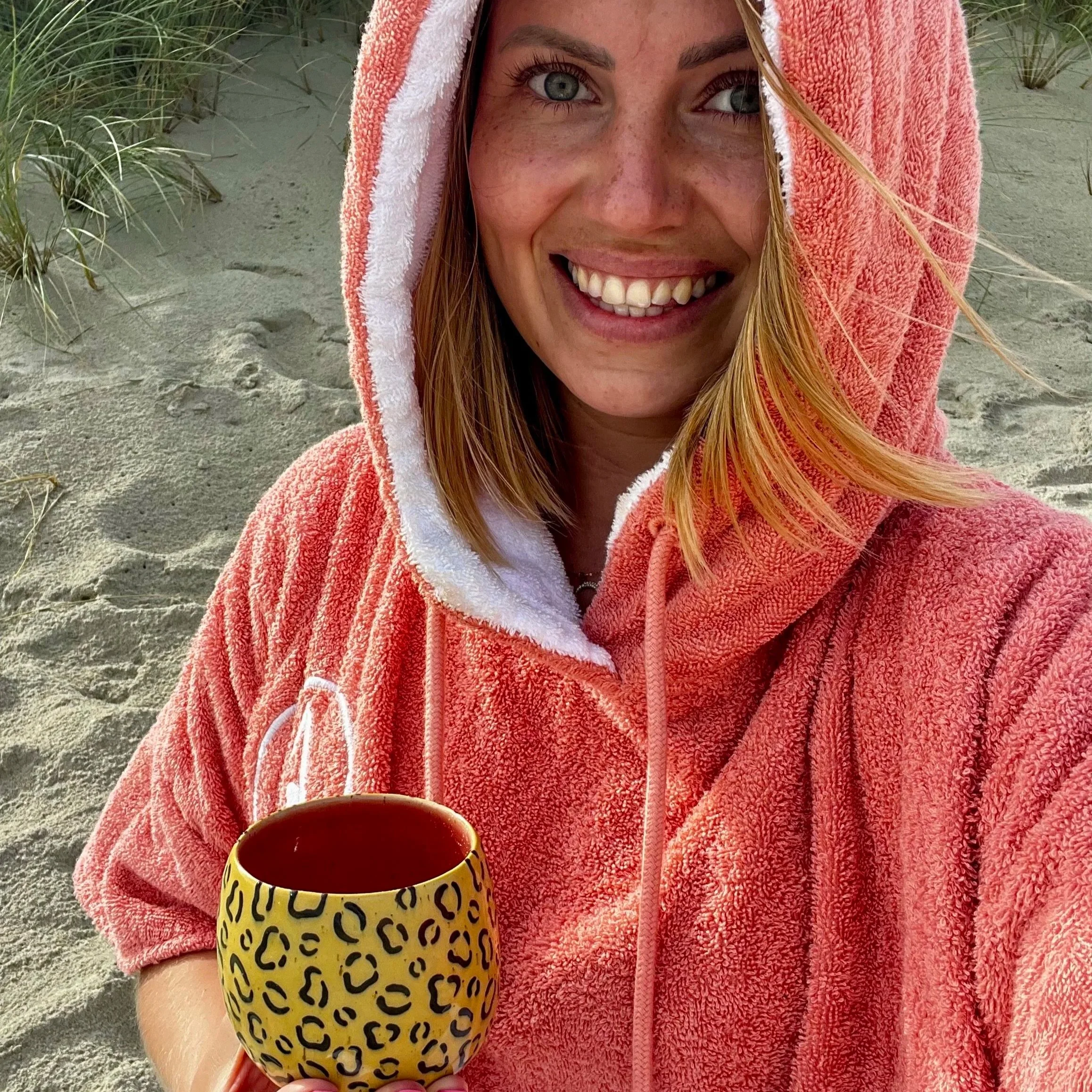 Eine lachende junge Frau mit blonden Haaren trägt einen pinken Surfponcho und hält eine gelbe Tasse mit schwarzem Leo-Muster in der Hand, sitzt am Strand mit Sand und Dünen im Hintergrund.