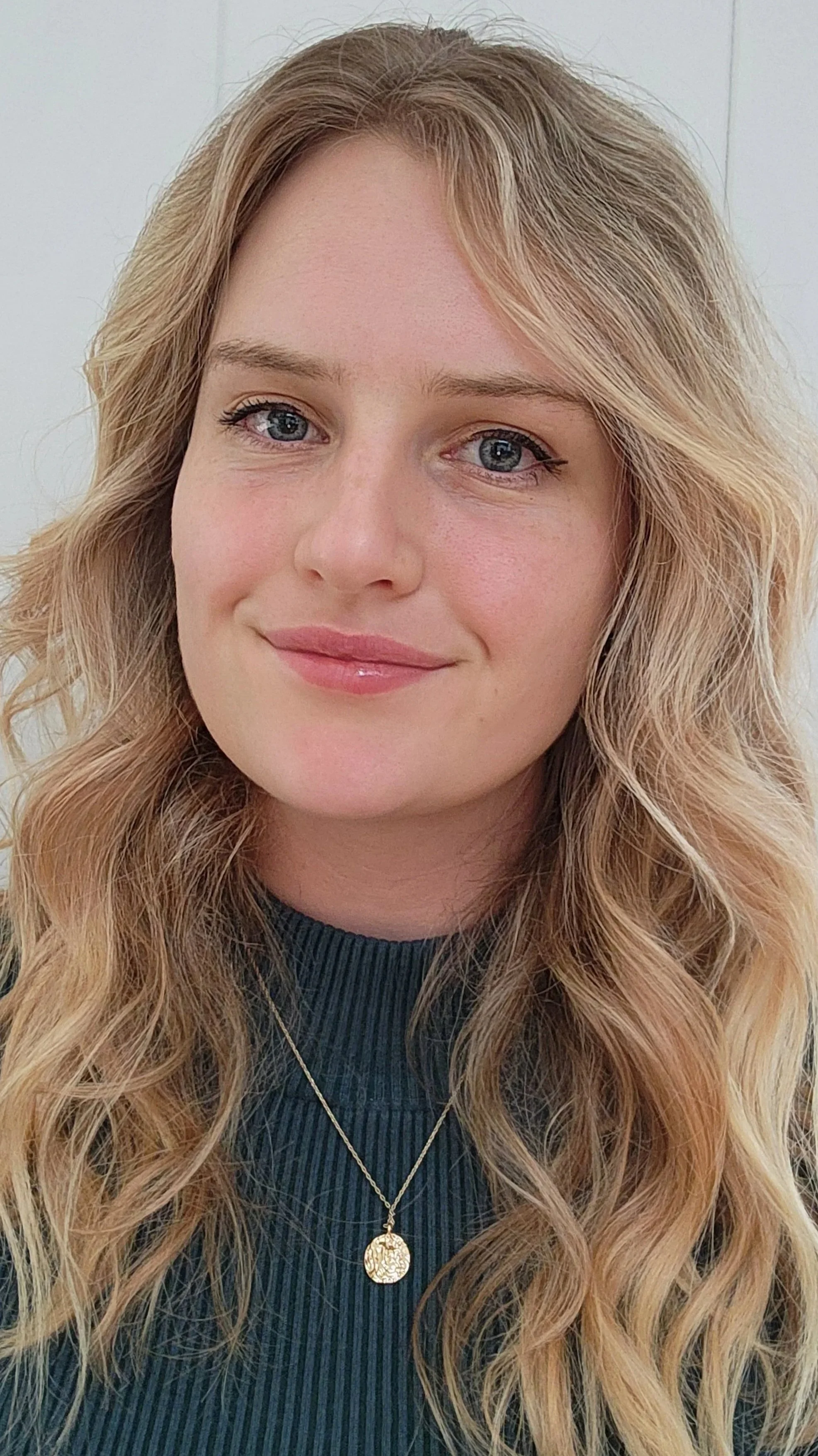 Elly Hanrahan; Close-up of a woman with blonde, wavy hair wearing a green ribbed top and a gold necklace with a circular pendant, smiling softly.