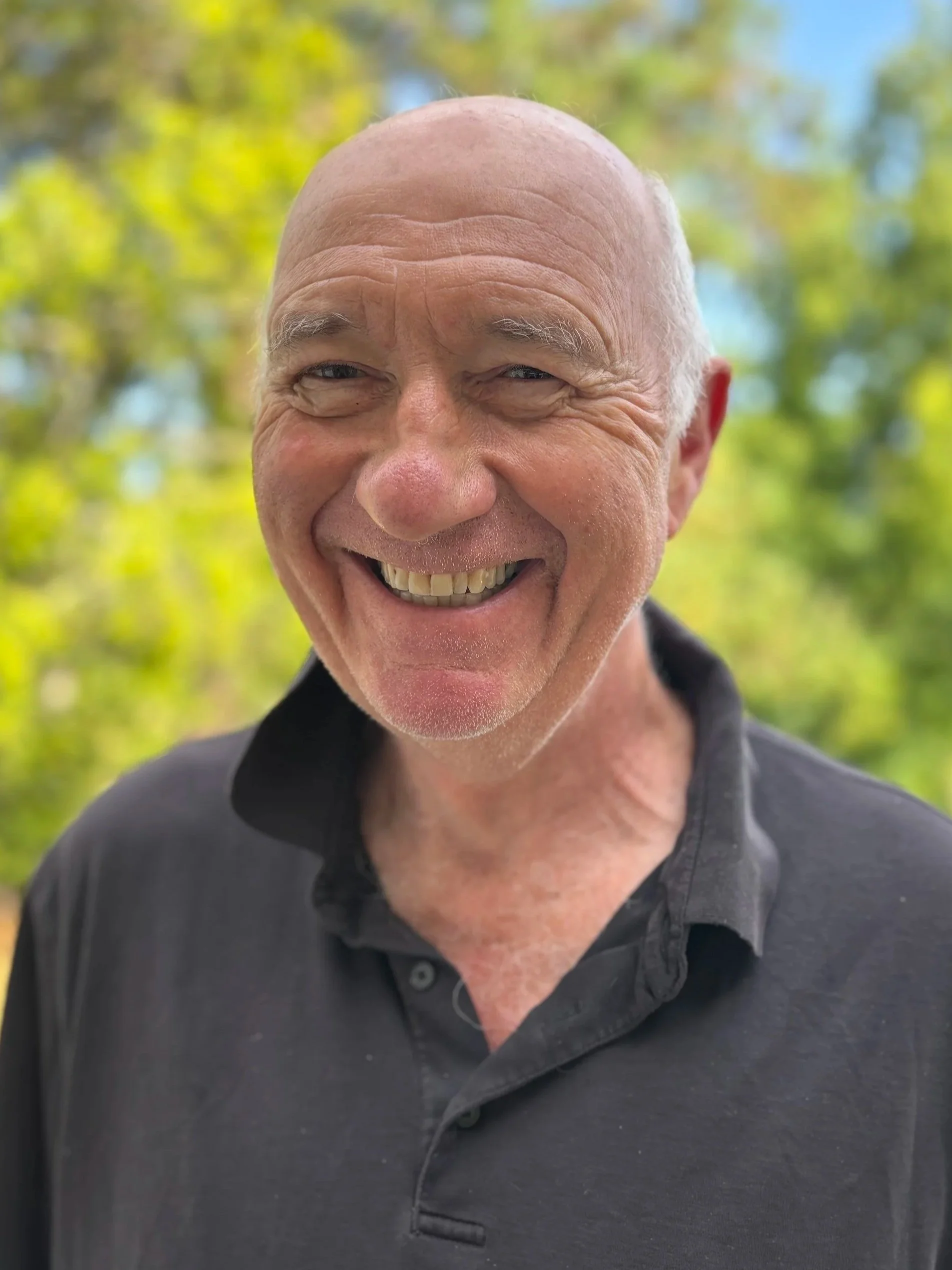 Jonathan Hamer; A smiling bald man outdoors with a blurred green and yellow background, wearing a black collared shirt.