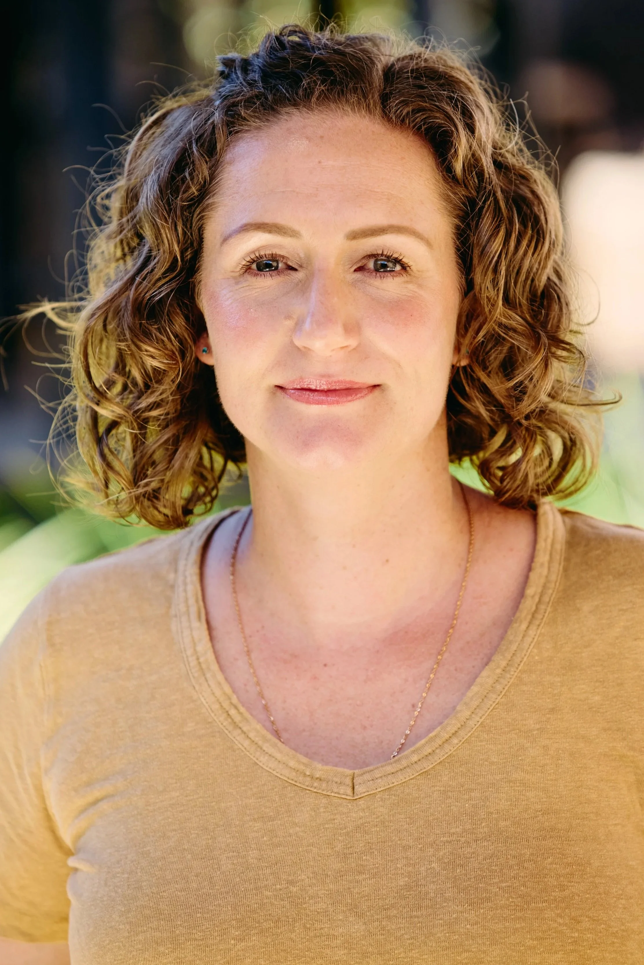 Close-up of a woman with curly brown hair, wearing a beige t-shirt and a delicate gold necklace, smiling outdoors with blurred background.