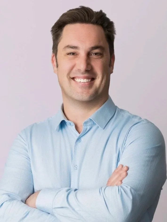 A smiling man with crossed arms, wearing a light blue dress shirt, against a plain light background.