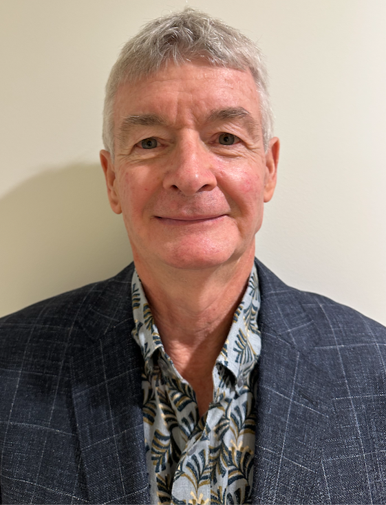 A middle-aged man with short gray hair, wearing a dark plaid blazer over a patterned shirt, smiling for a photo against a plain off-white wall.