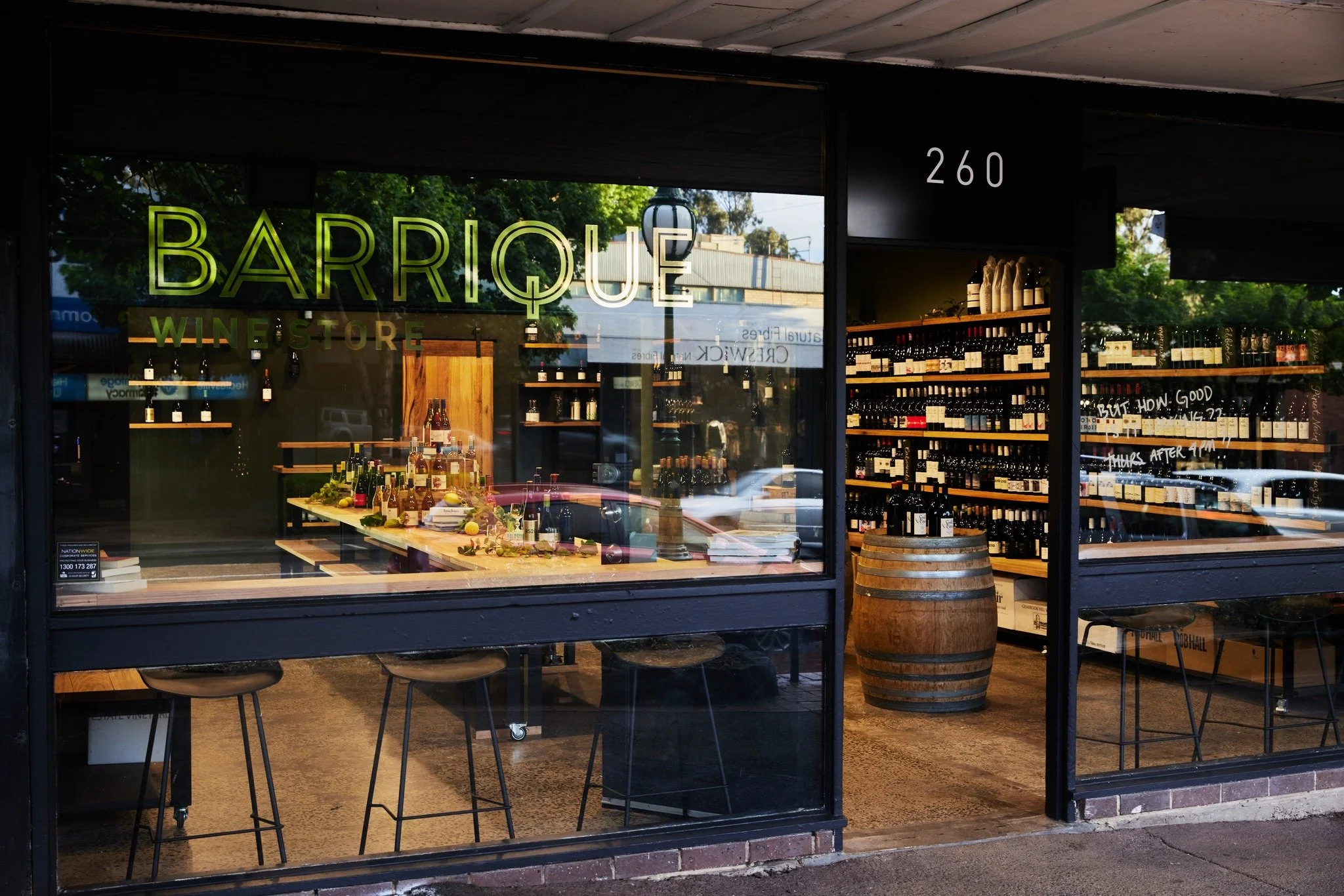 Front view of a wine store named 'Barrique' with a large window display showing wine bottles, a wooden barrel, tables with bottles and decorations inside, and a street outside.