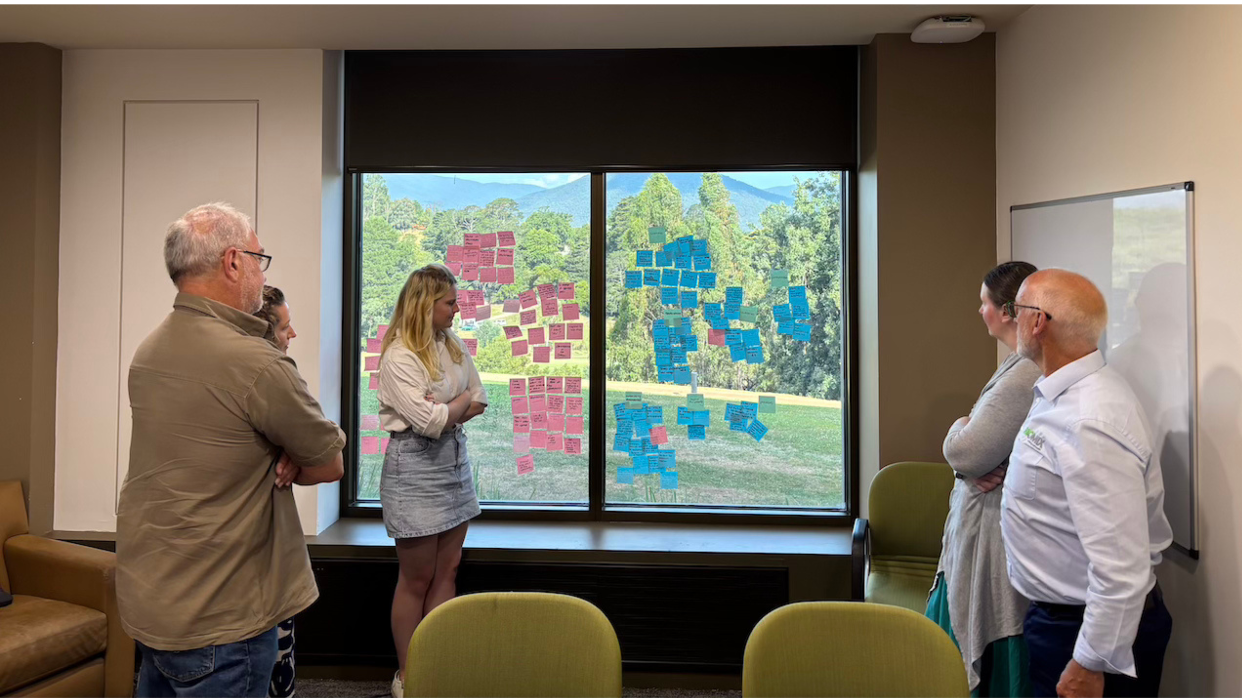 Group of five people in a conference room looking at colorful sticky notes on a window, with a scenic landscape of trees and mountains outside.