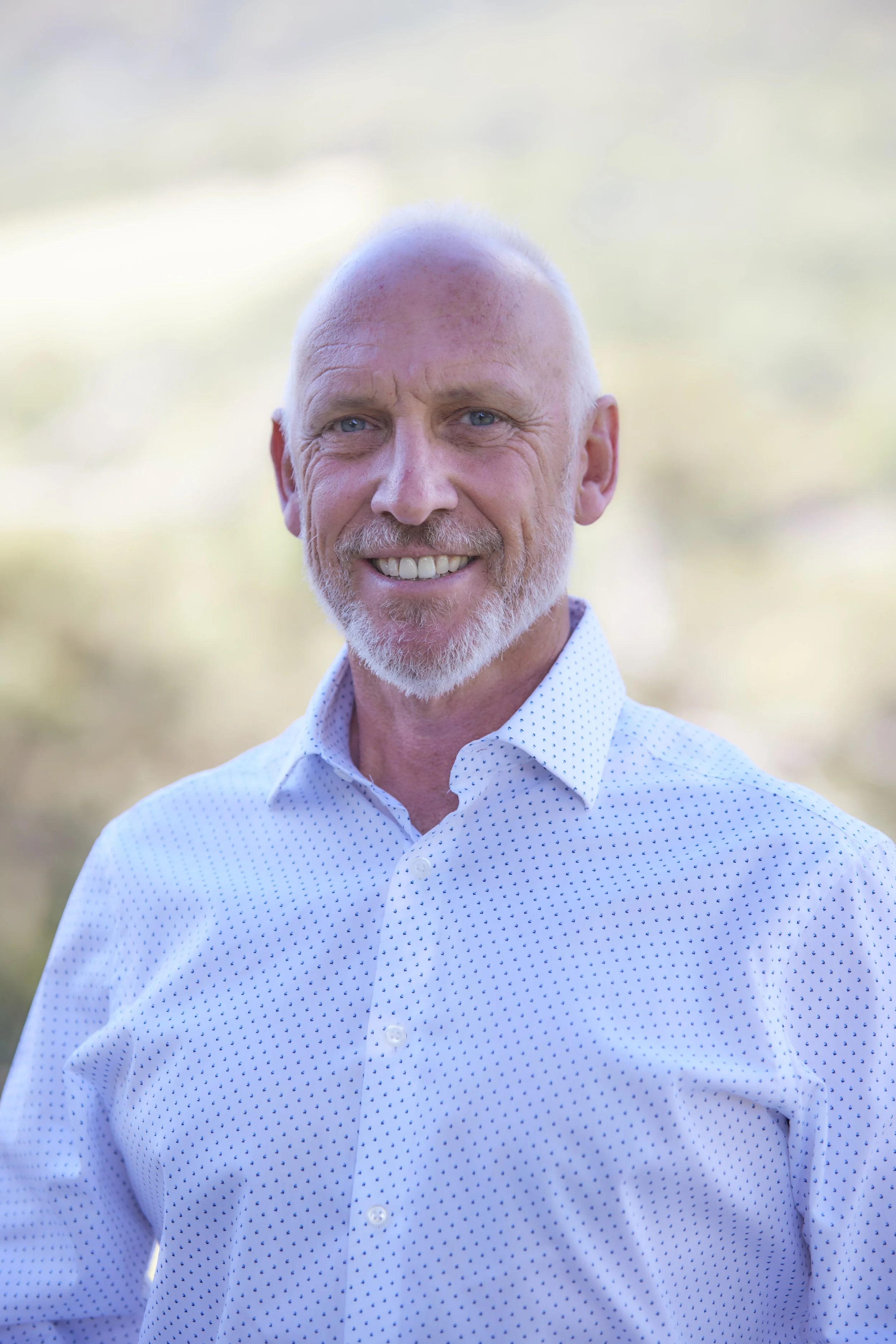 Michael Robertson; A smiling middle-aged man with a white beard and short white hair, wearing a white polka dot shirt, standing outdoors with a blurred natural background.