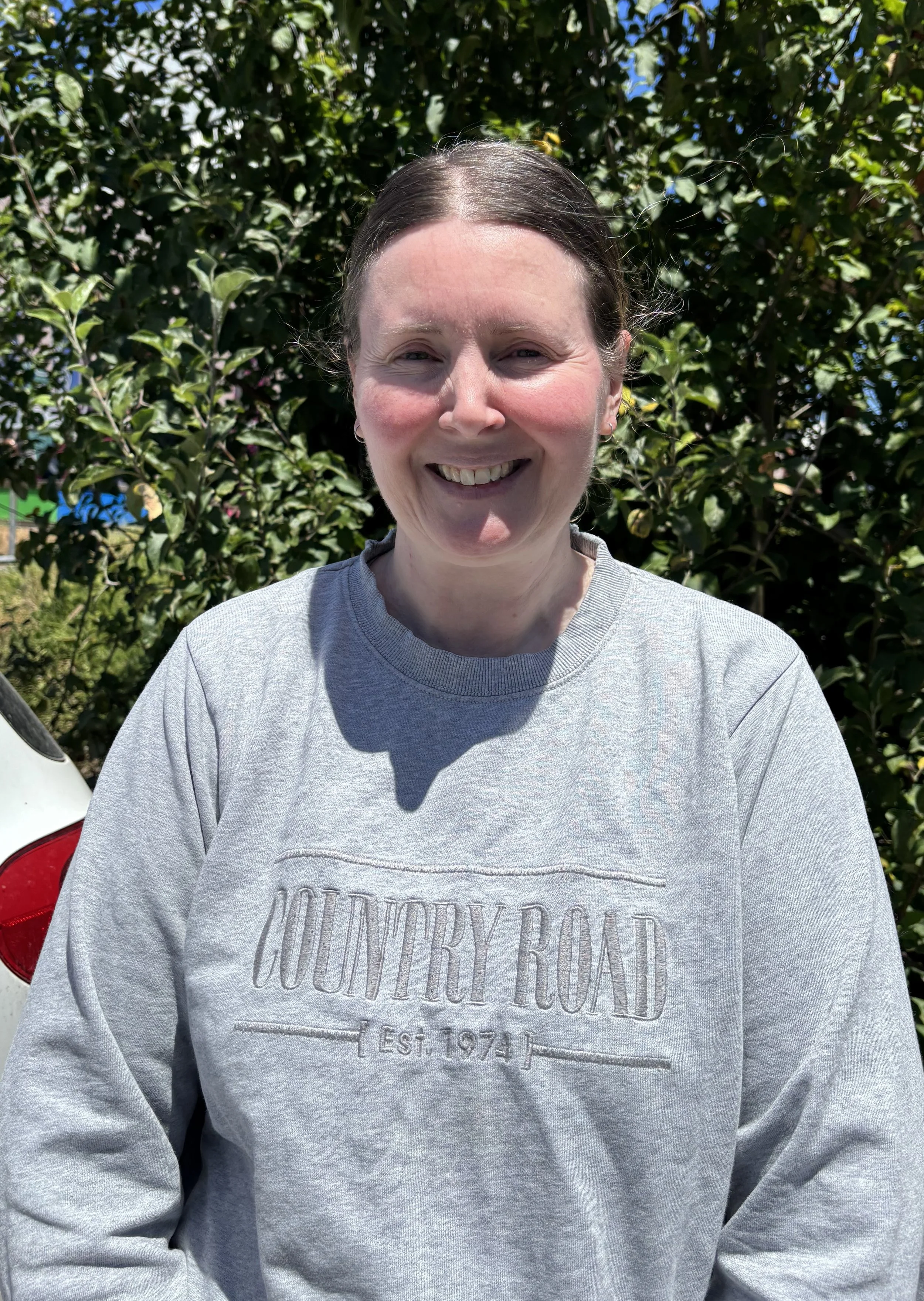 Joanne Fuller; A smiling woman with dark brown hair pulled back, wearing a gray sweatshirt with the text 'COUNTRY ROAD EST. 1974,' standing outdoors in front of green foliage.