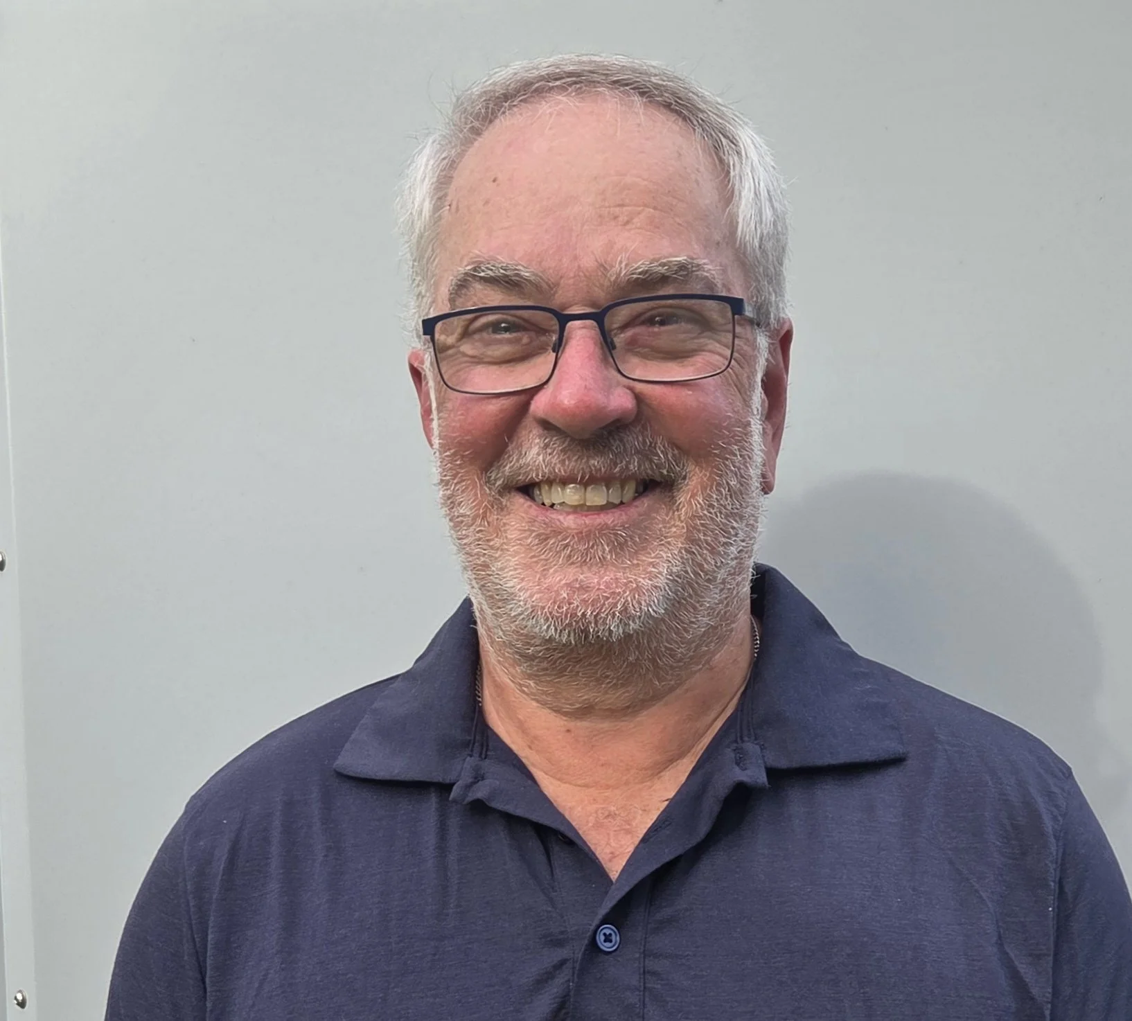 Greg Whimp; Smiling middle-aged man with glasses, short gray hair, a beard, wearing a navy blue polo shirt, standing against a plain light-colored wall.